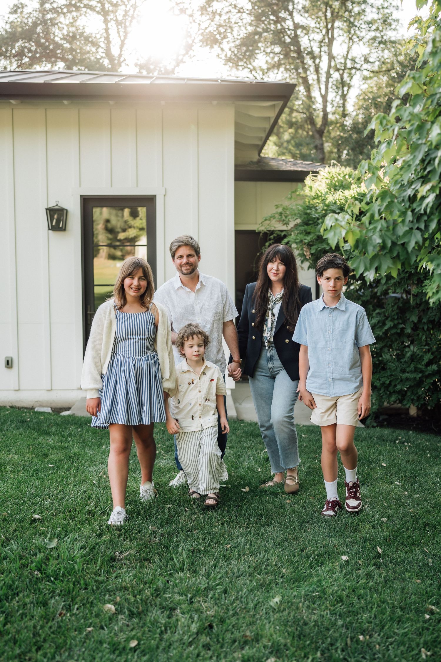 Family of five posing together on green lawn in front of white house, surrounded by lush trees on sunny day.
