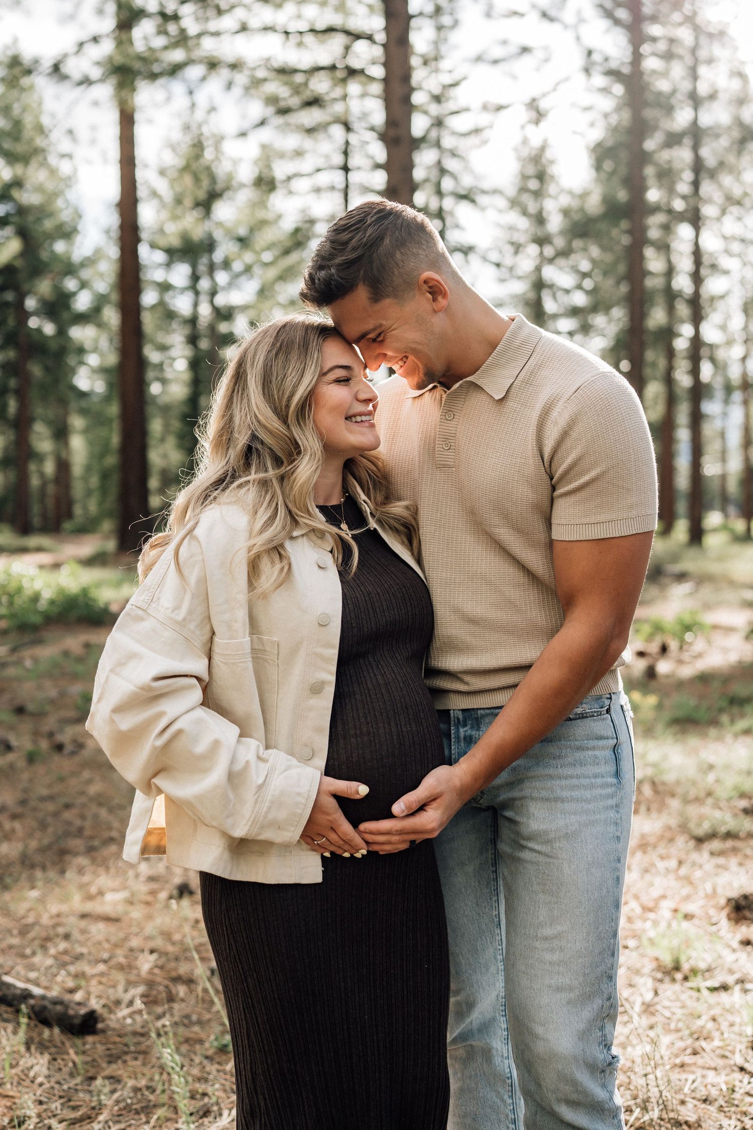 Pregnant couple embracing in a forest, woman in black dress and cream jacket, man in beige polo and jeans.