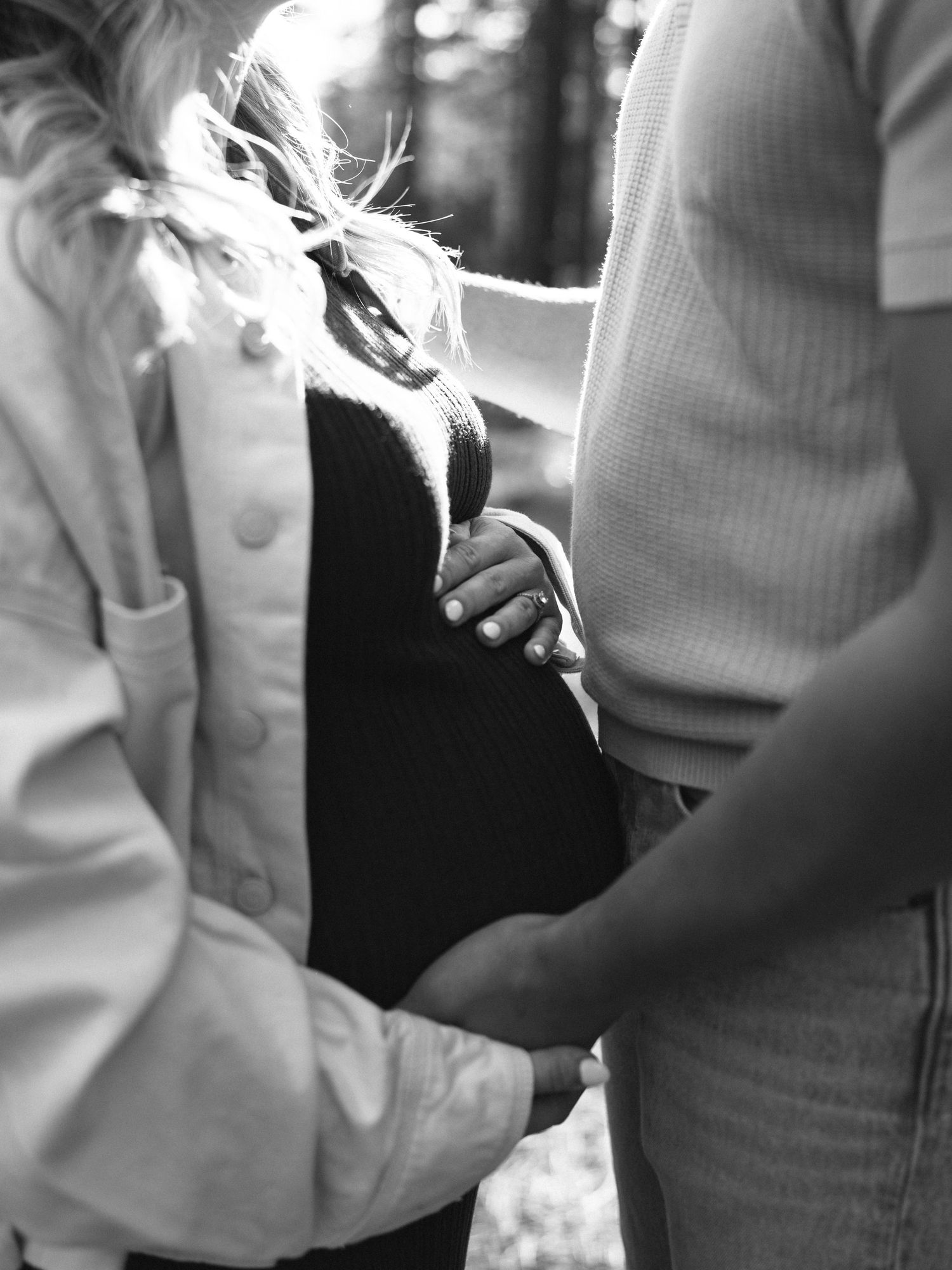 Black and white photo of a pregnant woman holding her baby bump with a child touching her belly outdoors.