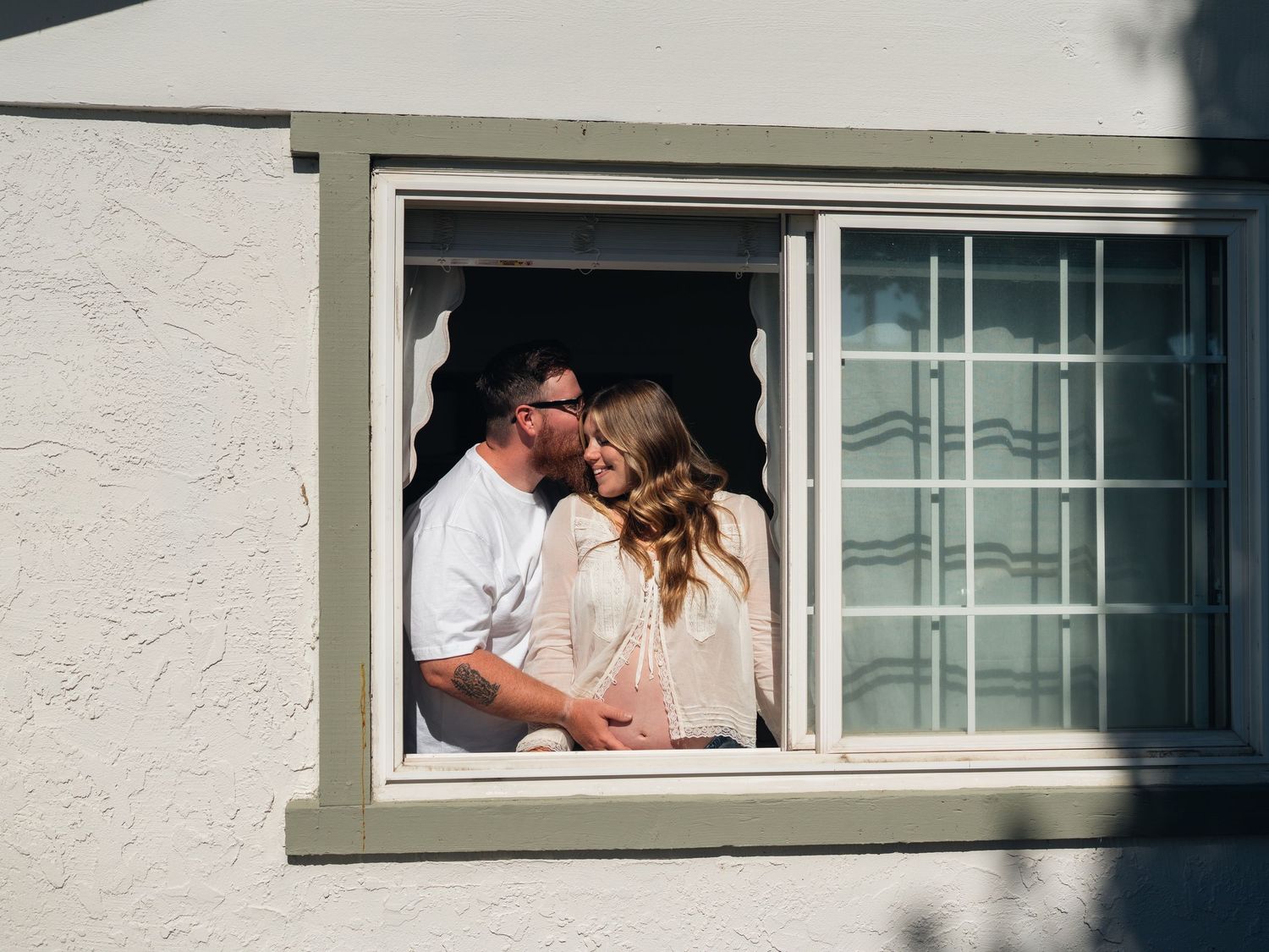 Couple sharing a romantic kiss framed in an open window of a white stucco building.