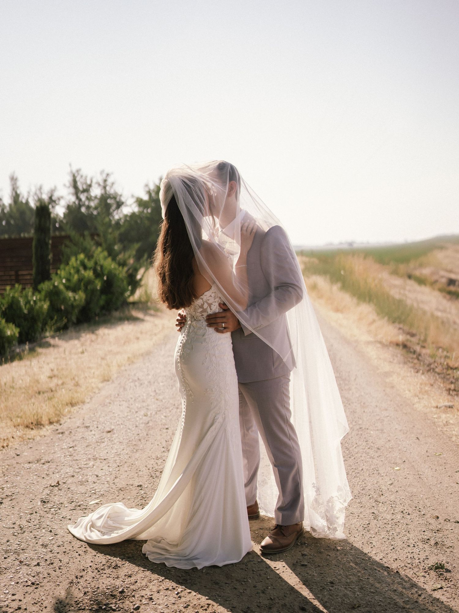 Romantic couple in wedding attire walking along a sunlit dirt path.