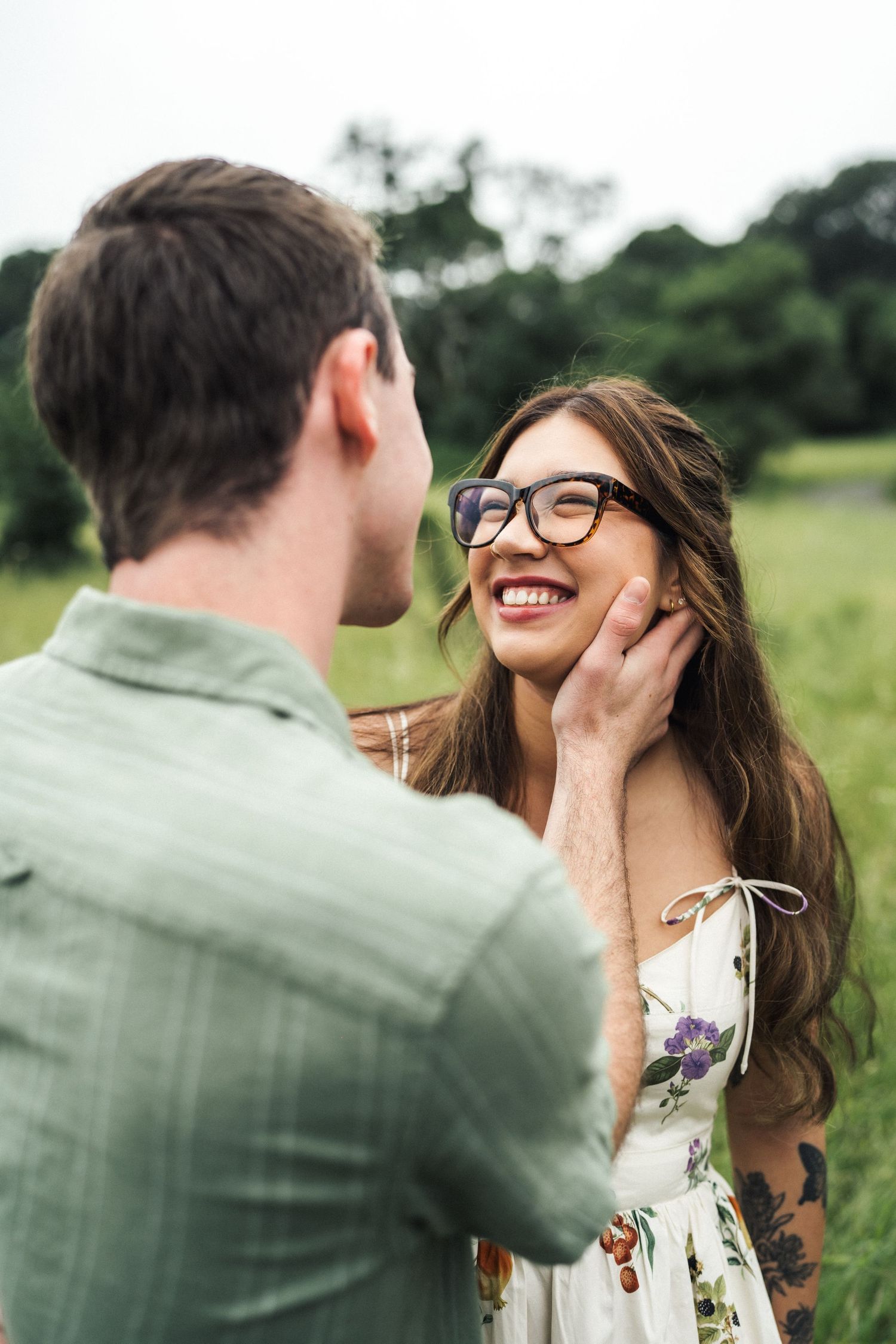 A smiling woman in floral dress and glasses laughs as a man in green shirt gently touches her face in a green field.