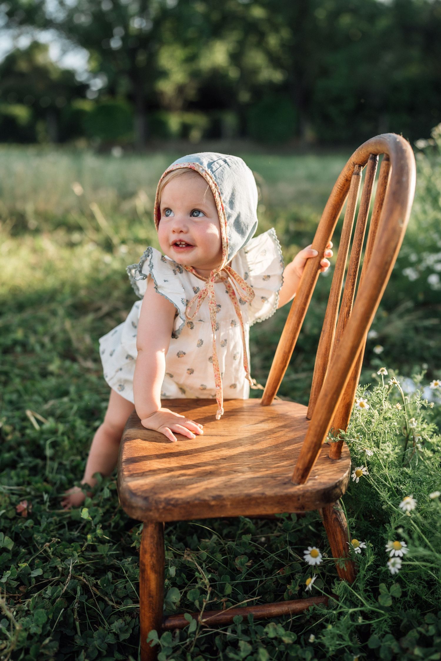 A baby in a floral romper and bonnet sits on a wooden chair in a sunny green field with daisies.