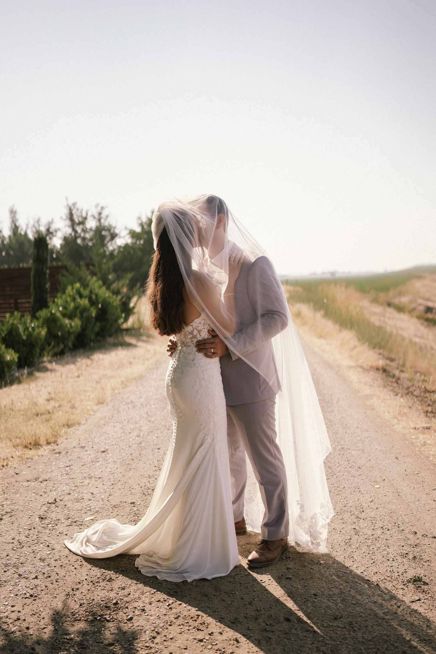 Wedding couple sharing intimate moment on dirt road with flowing veil in breeze.