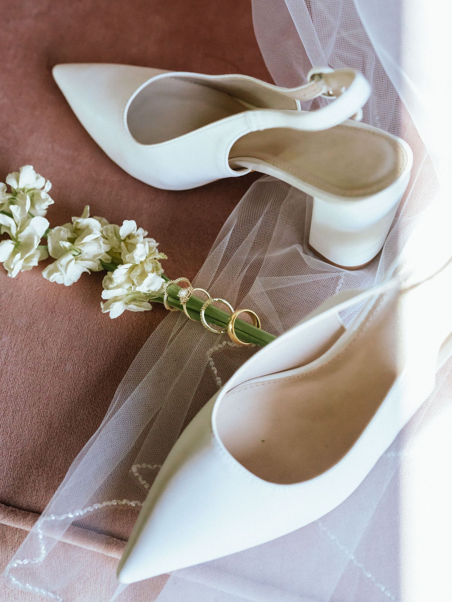 White bridal heels with delicate flowers and wedding rings arranged on brown fabric.
