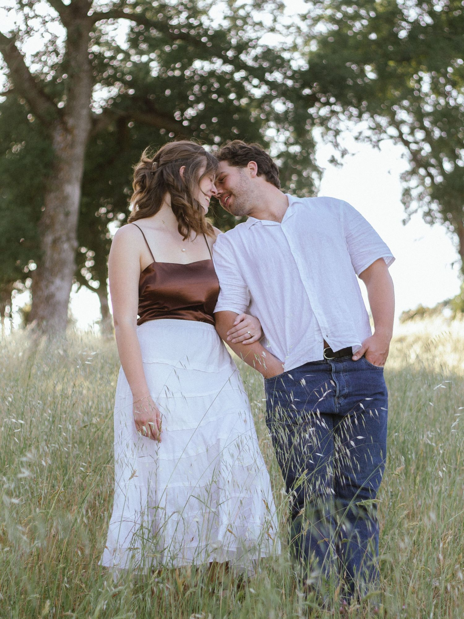 Romantic couple in white and denim embrace while standing in a tall grass meadow at sunset.