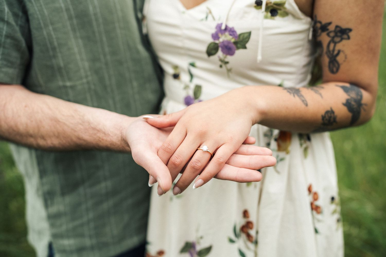 Couple holding hands showing engagement ring, woman with floral dress and tattoo, man in green shirt outdoors.
