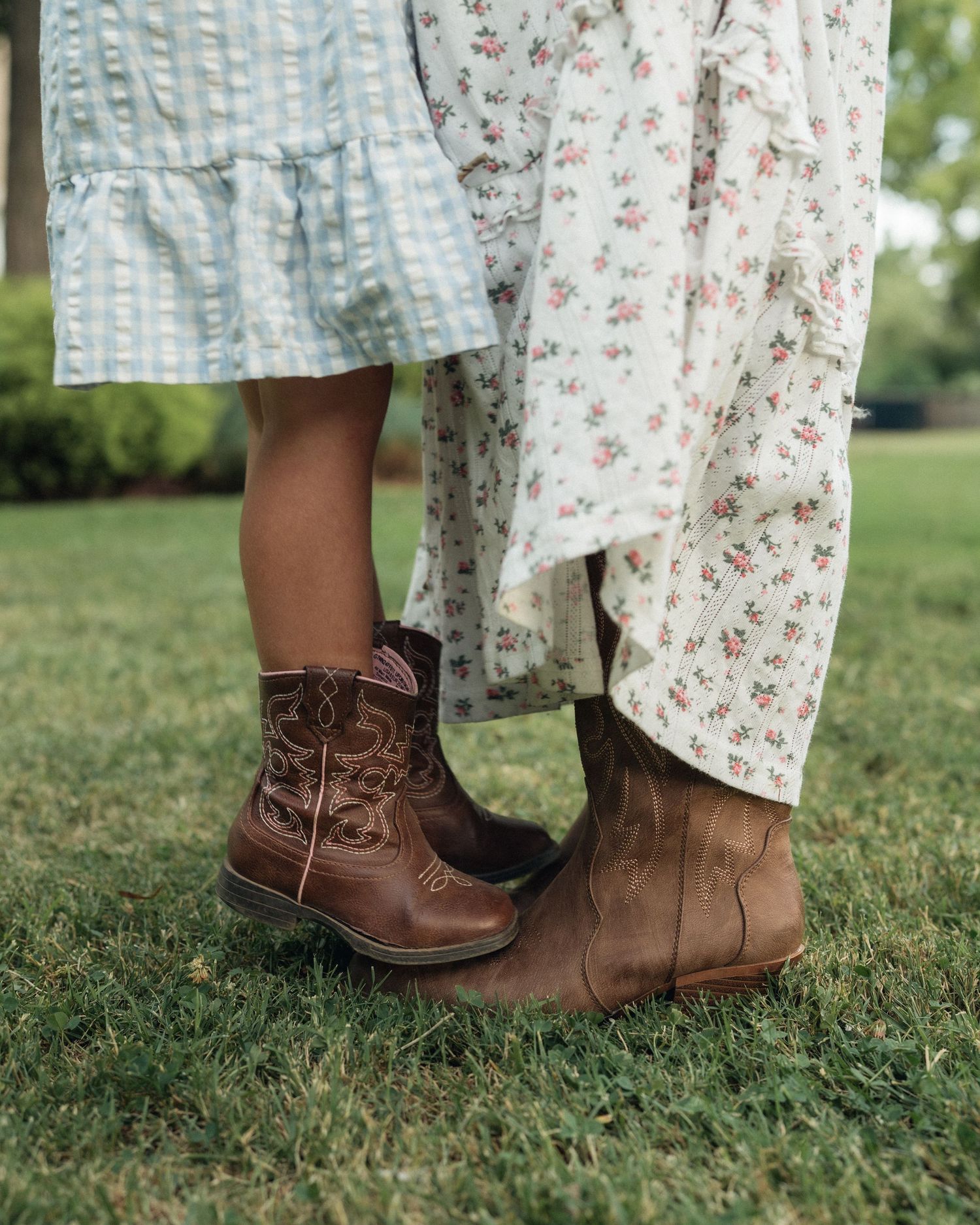 Two people in floral dresses wearing brown cowboy boots standing on green grass.