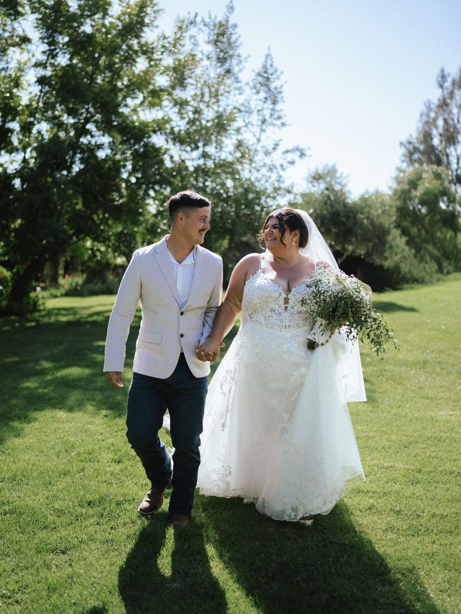 A couple in wedding attire walk together on a grassy lawn during a sunny outdoor celebration.