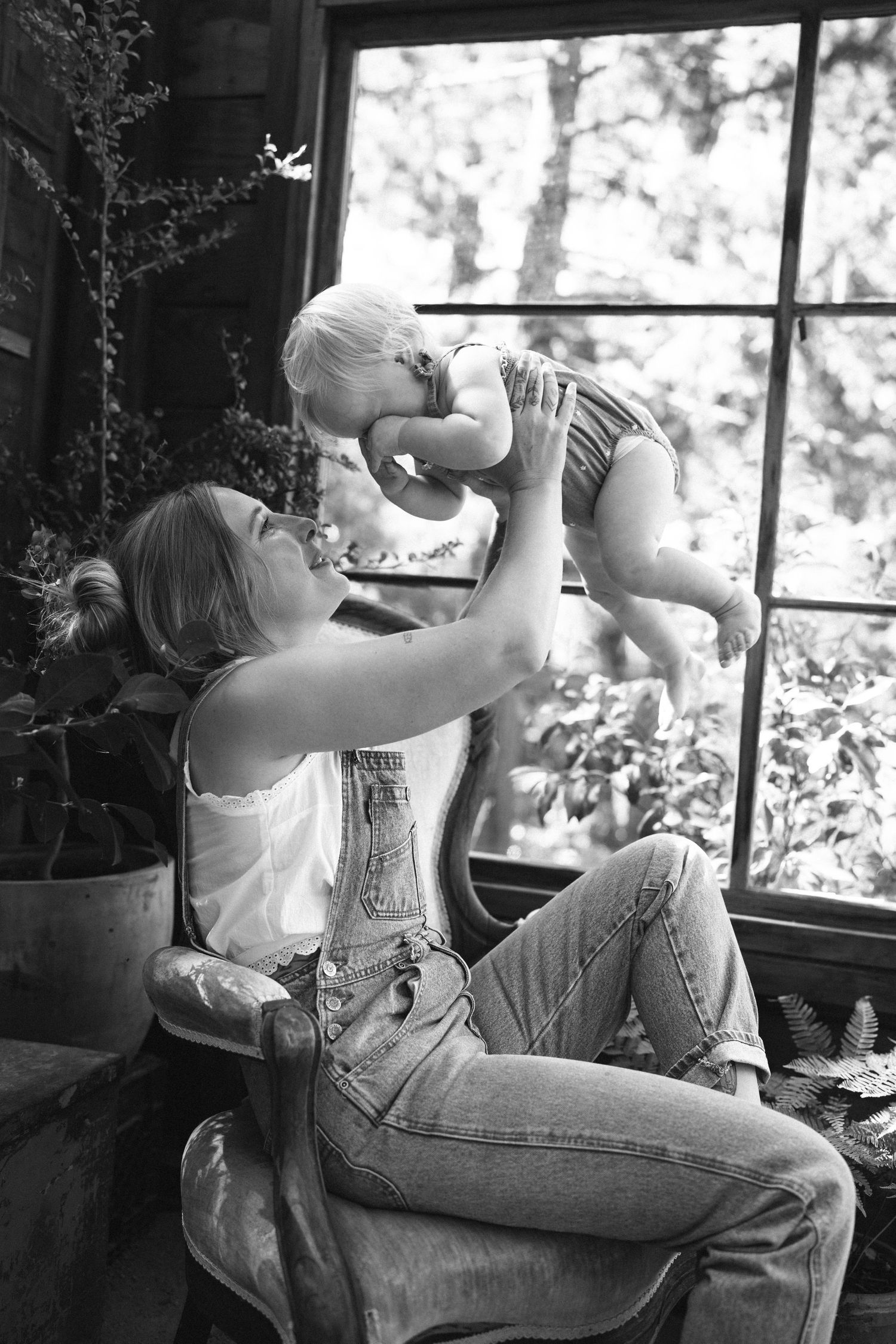 Black and white photo of a mother lifting her baby in the air while sitting in a chair near a large window with plants.