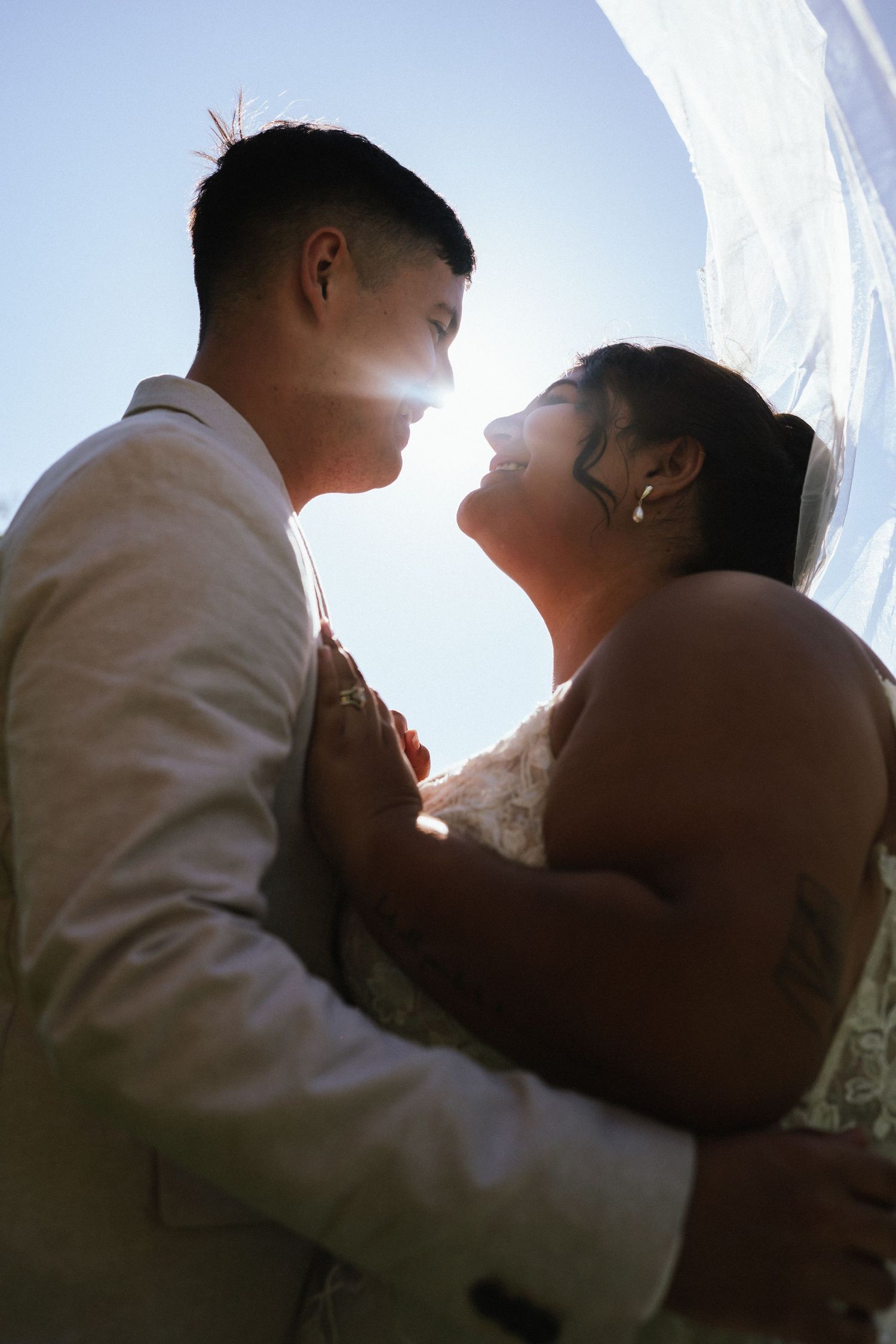 Backlit wedding couple embracing under bright sky with veil flowing.