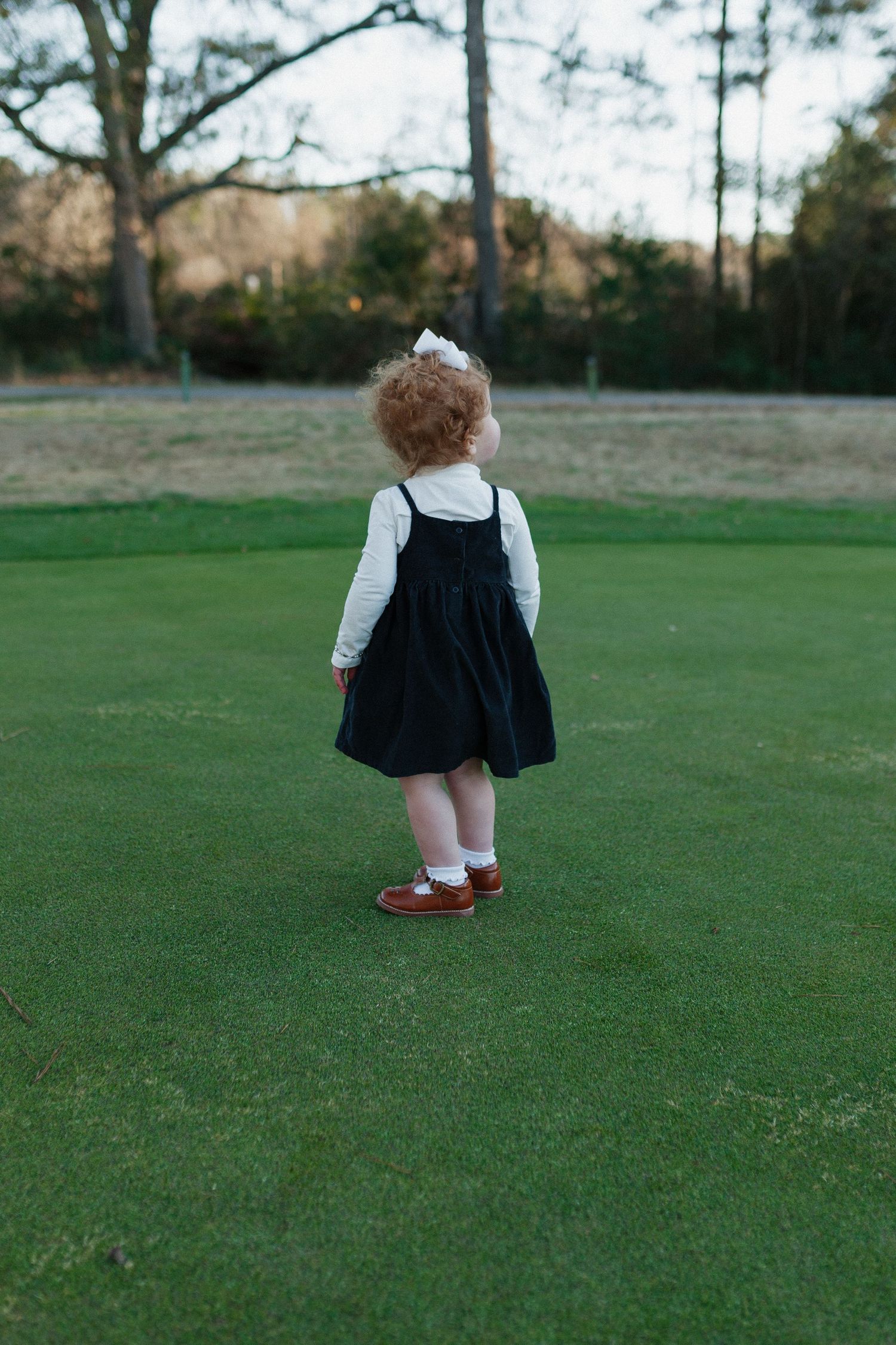 Child in black pinafore dress walking across a green lawn with trees in the background. Alabama photographer; South Alabama photographer