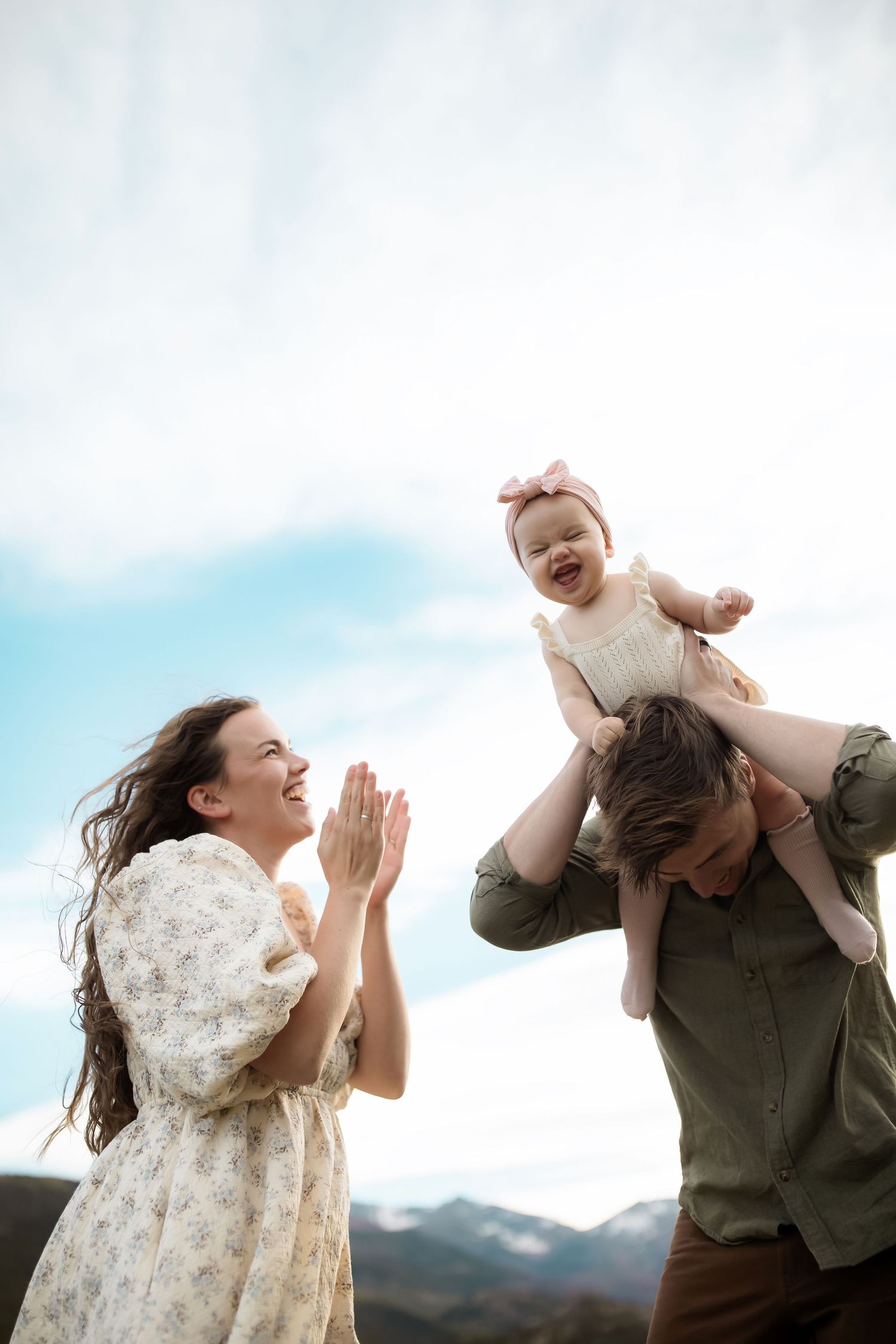 Happy family outdoors, mother clapping as father lifts laughing baby girl against blue sky and mountain backdrop.