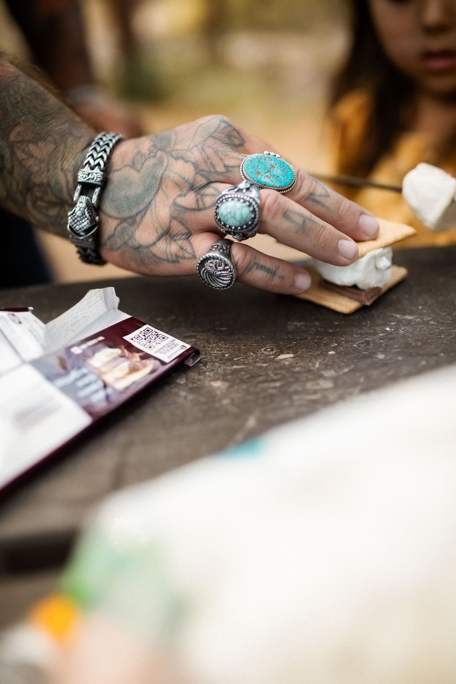 Tattooed hands with turquoise rings shown working with paper materials on dark surface.