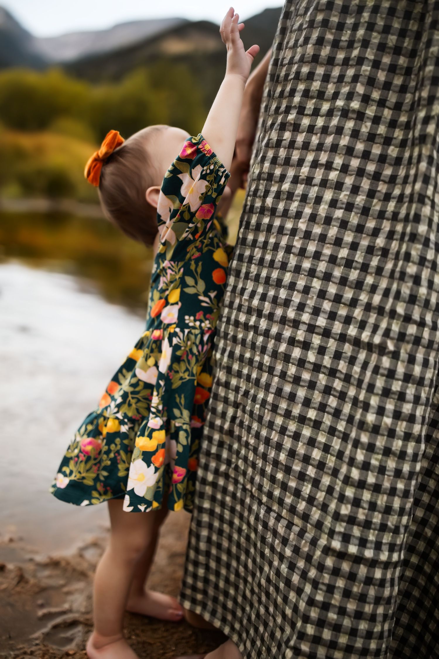 Small child in floral dress reaching up against black and white checkered fabric outdoors by water.