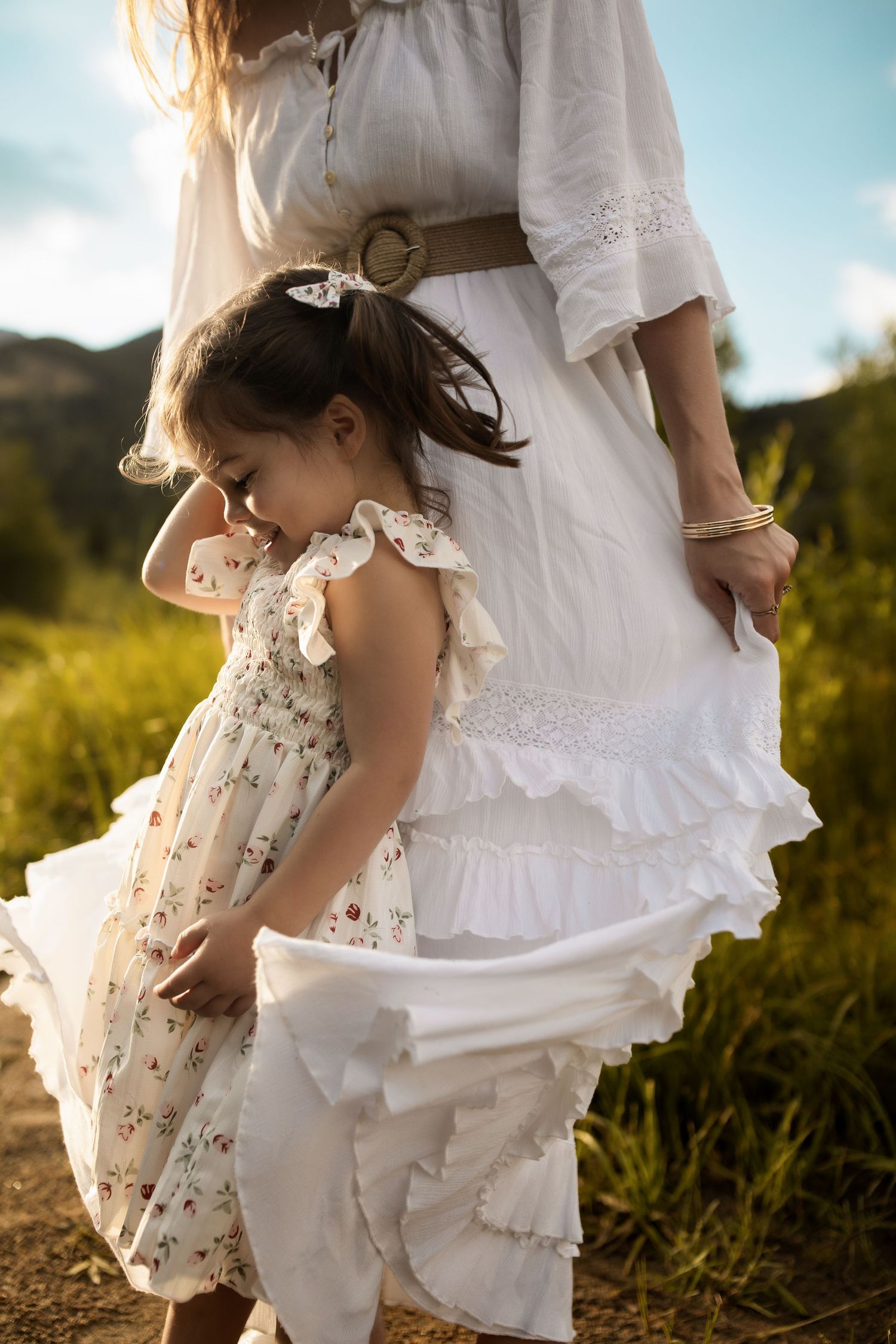 A young girl in a floral dress stands beside a woman in a white boho dress outdoors in a sunny field.