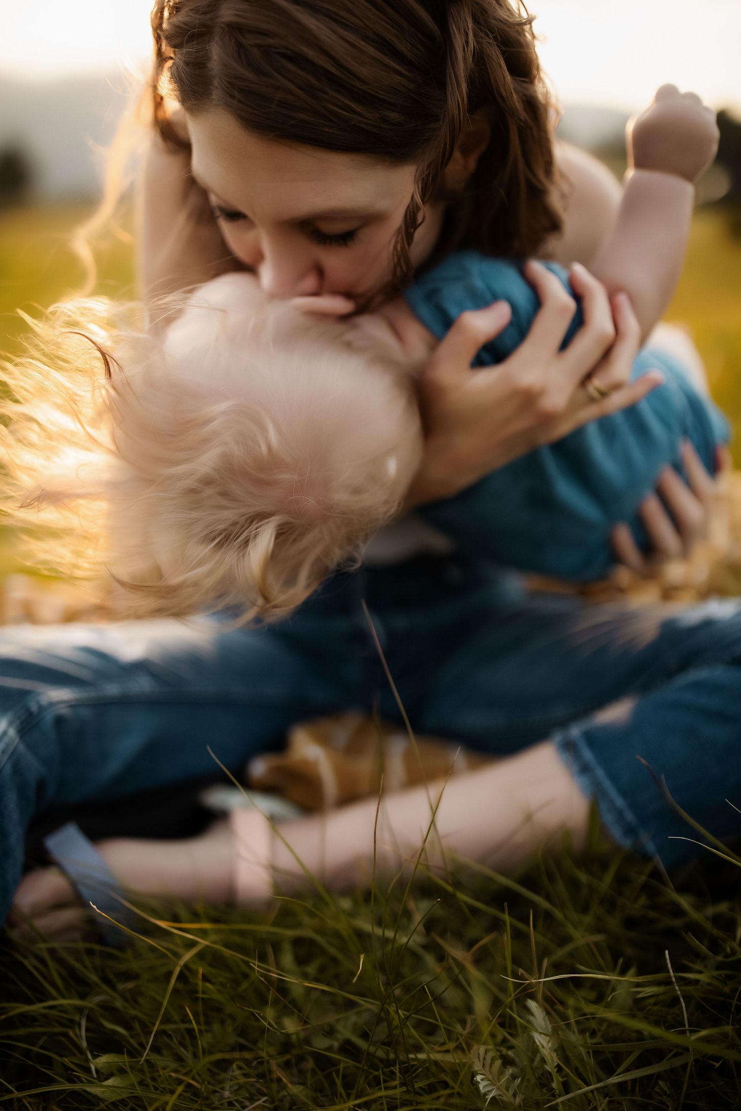 A mother tenderly kisses her young blonde toddler while sitting together in a sunlit green meadow.