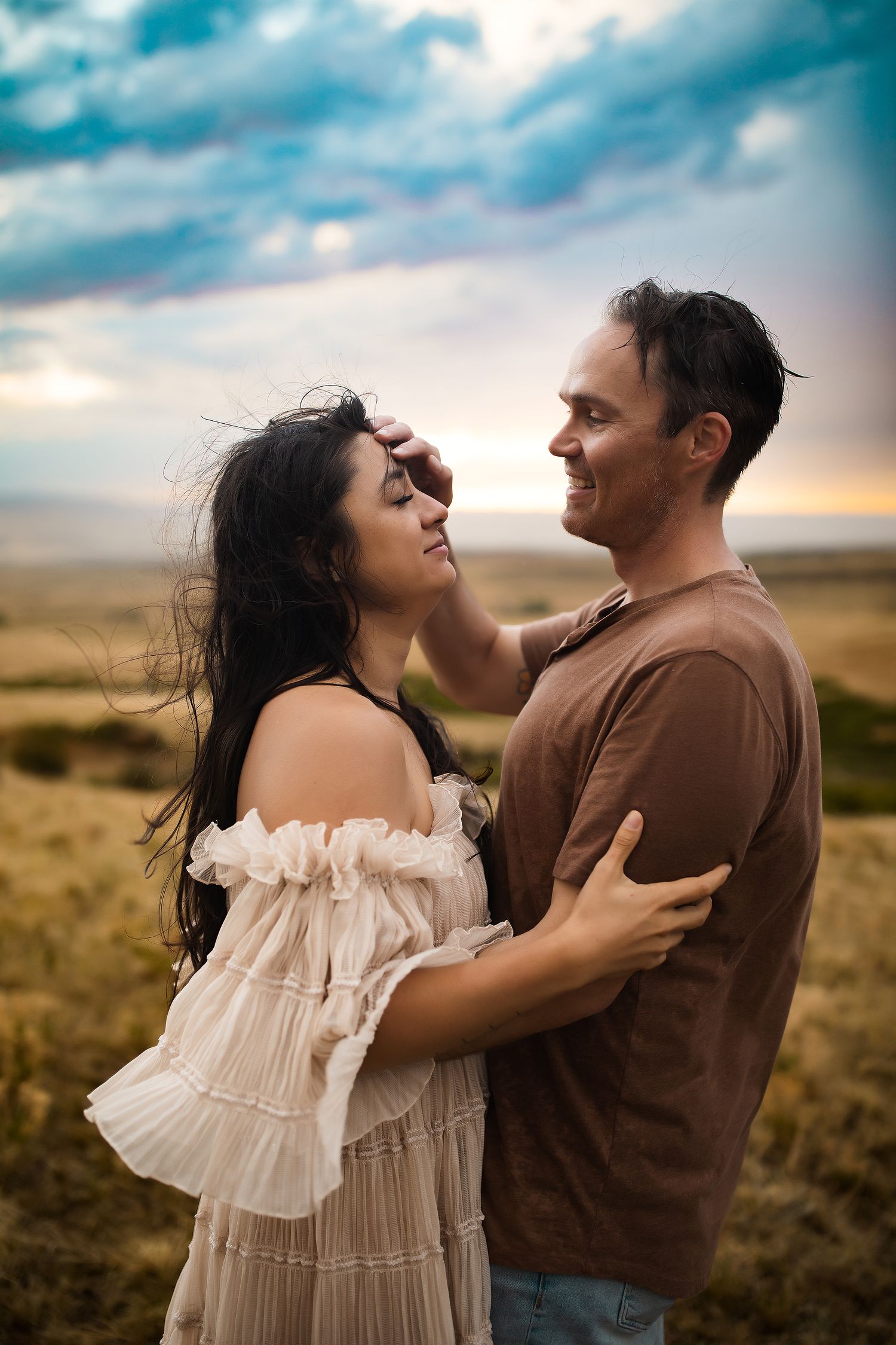 A man and a woman embrace in a romantic moment against a scenic backdrop of a cloudy sky and open field.