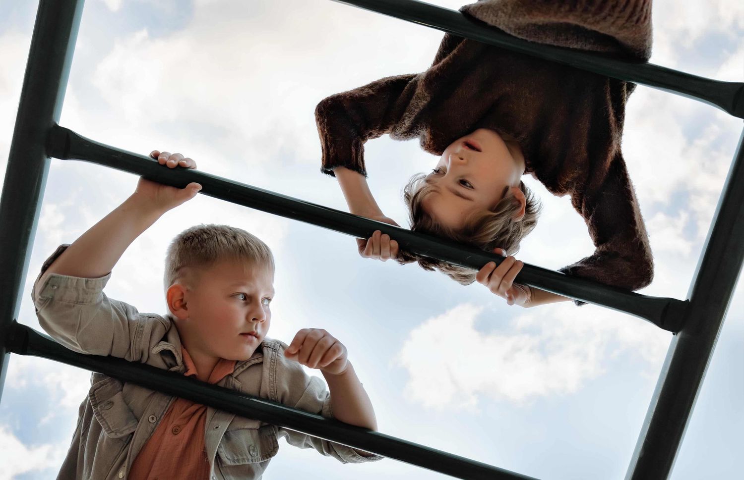 Two children playing on monkey bars at a playground, one hanging upside down against a cloudy blue sky.