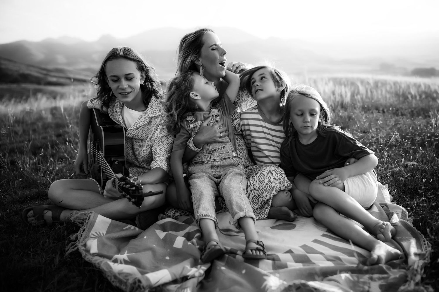 A group relaxes together on a picnic blanket in a sunny meadow with rolling hills in the background.