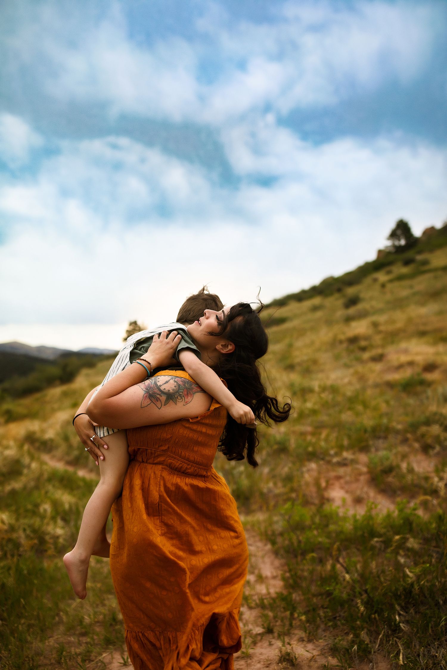 A tender embrace between two people on a grassy hillside, one wearing a flowing rust-colored dress.
