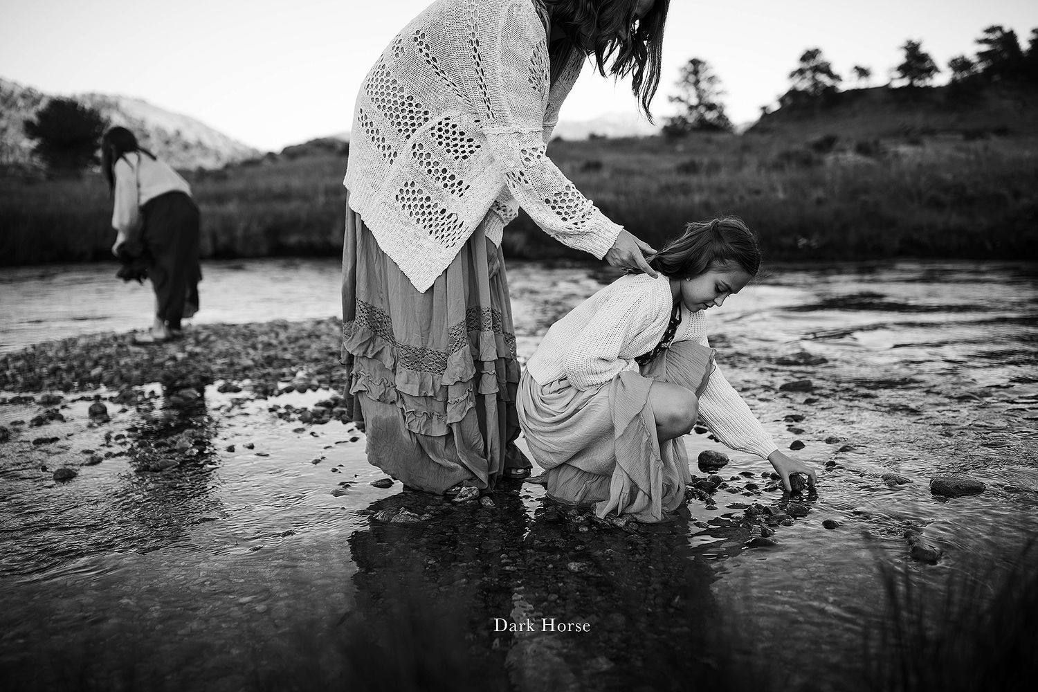 People in flowing dresses walk along rocky beach shoreline in black and white.