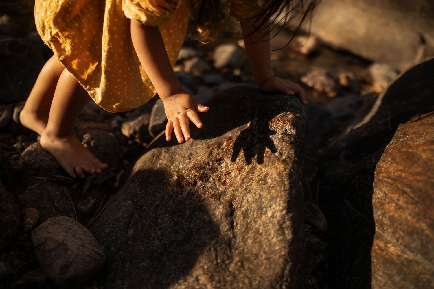 Small hands reach down to touch rough stone surfaces in warm golden sunlight creating dramatic shadows.