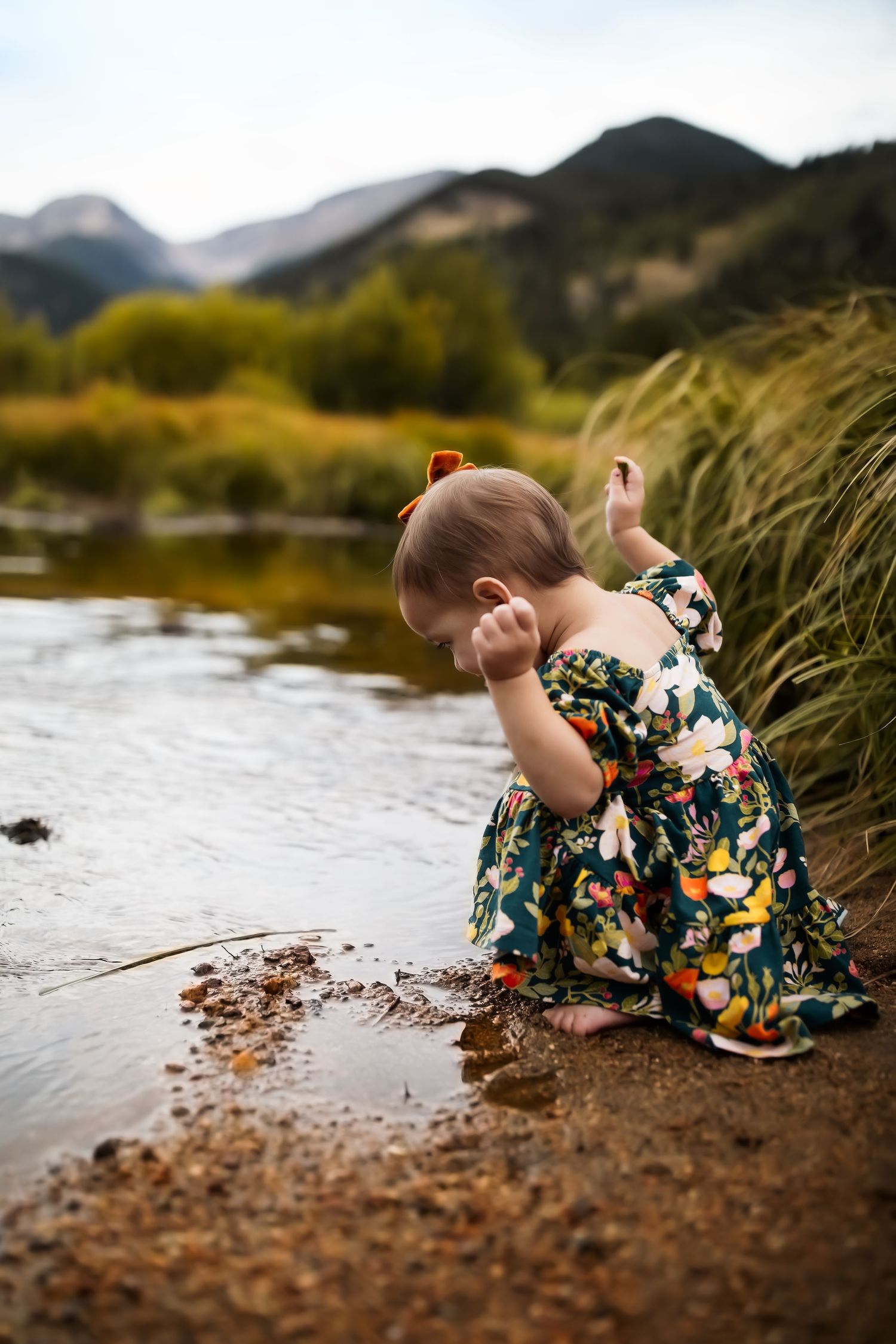 A child in a floral dress explores the riverbank while playing in the mud and water.