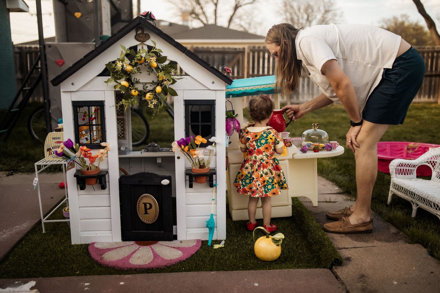 A mother and toddler girl in floral dress play outside a decorated white playhouse in a backyard setting.