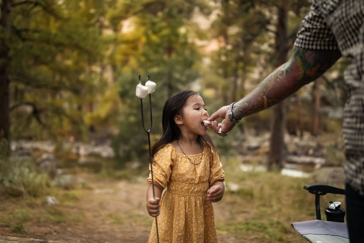 A child in a yellow dress holds white flowers while being offered a drink in an autumn forest setting.