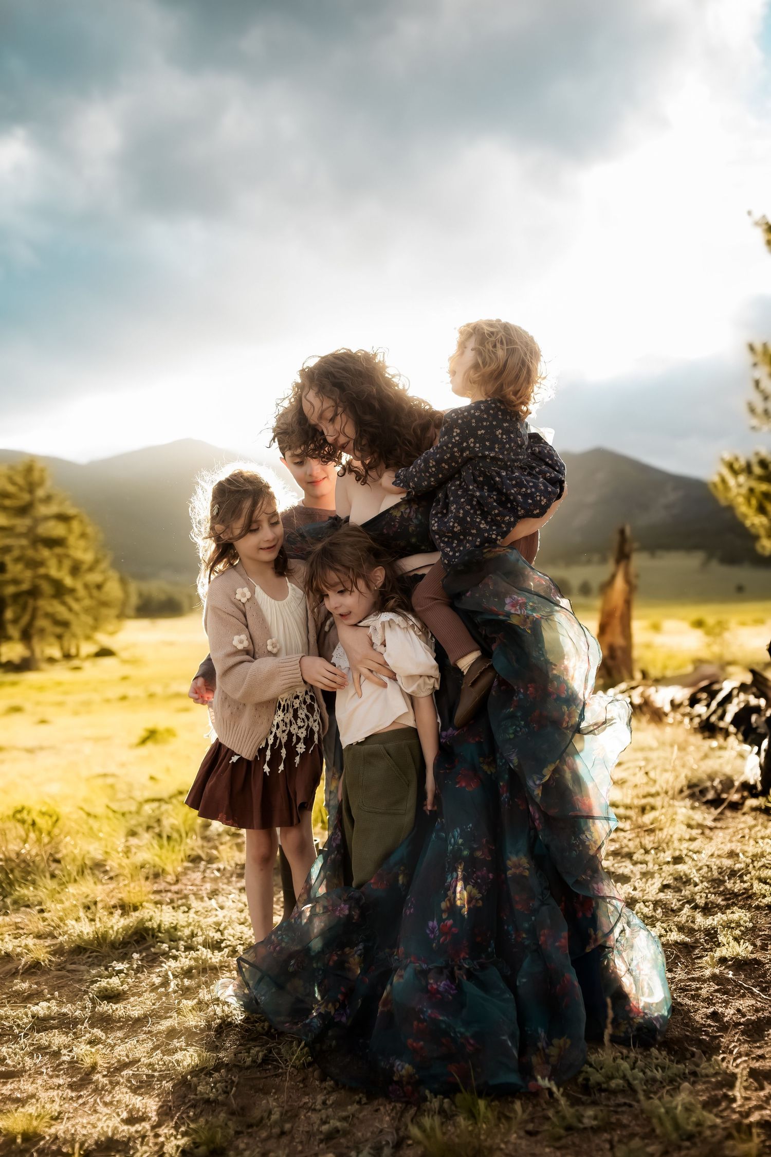 A mother in a floral skirt embraces her four young children in a sunlit mountain meadow.