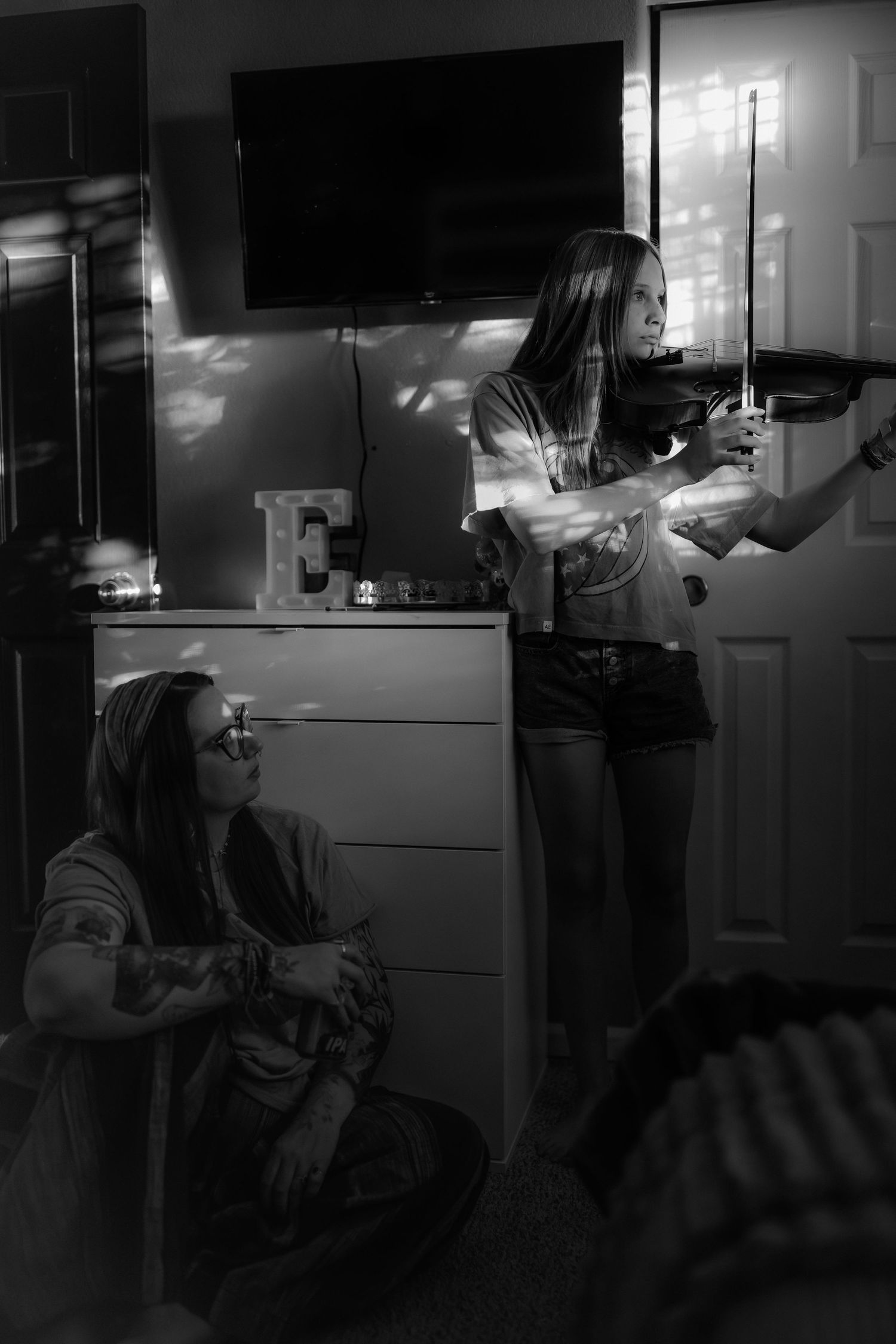 Black and white photo of a girl playing violin in a bedroom while another person sits on the floor watching.