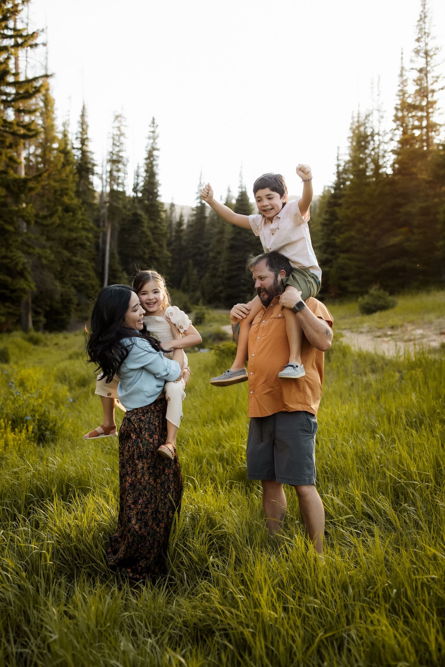Family of four enjoying a sunny outdoor photo session in a lush green mountain meadow surrounded by pine trees.