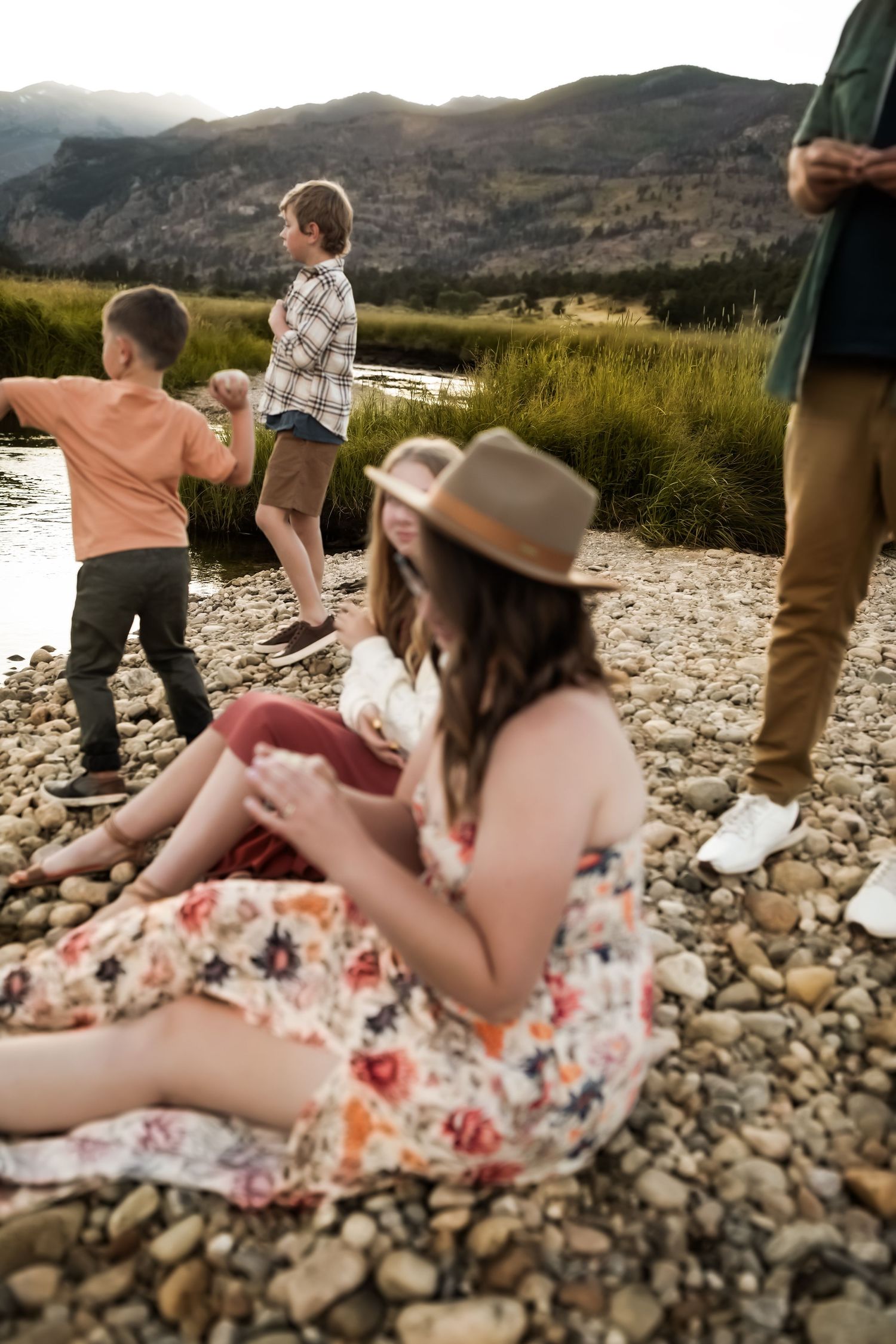 Family relaxing together on rocky riverbank with mountains in background.
