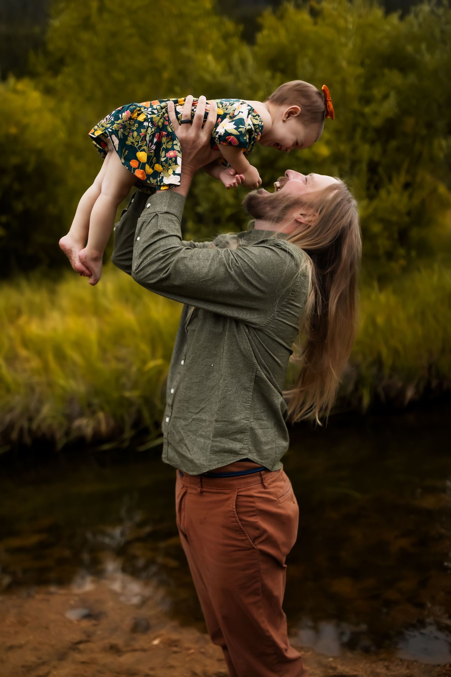 Joyful outdoor moment with floral-dressed toddler being lifted up by adult in olive shirt by riverside.