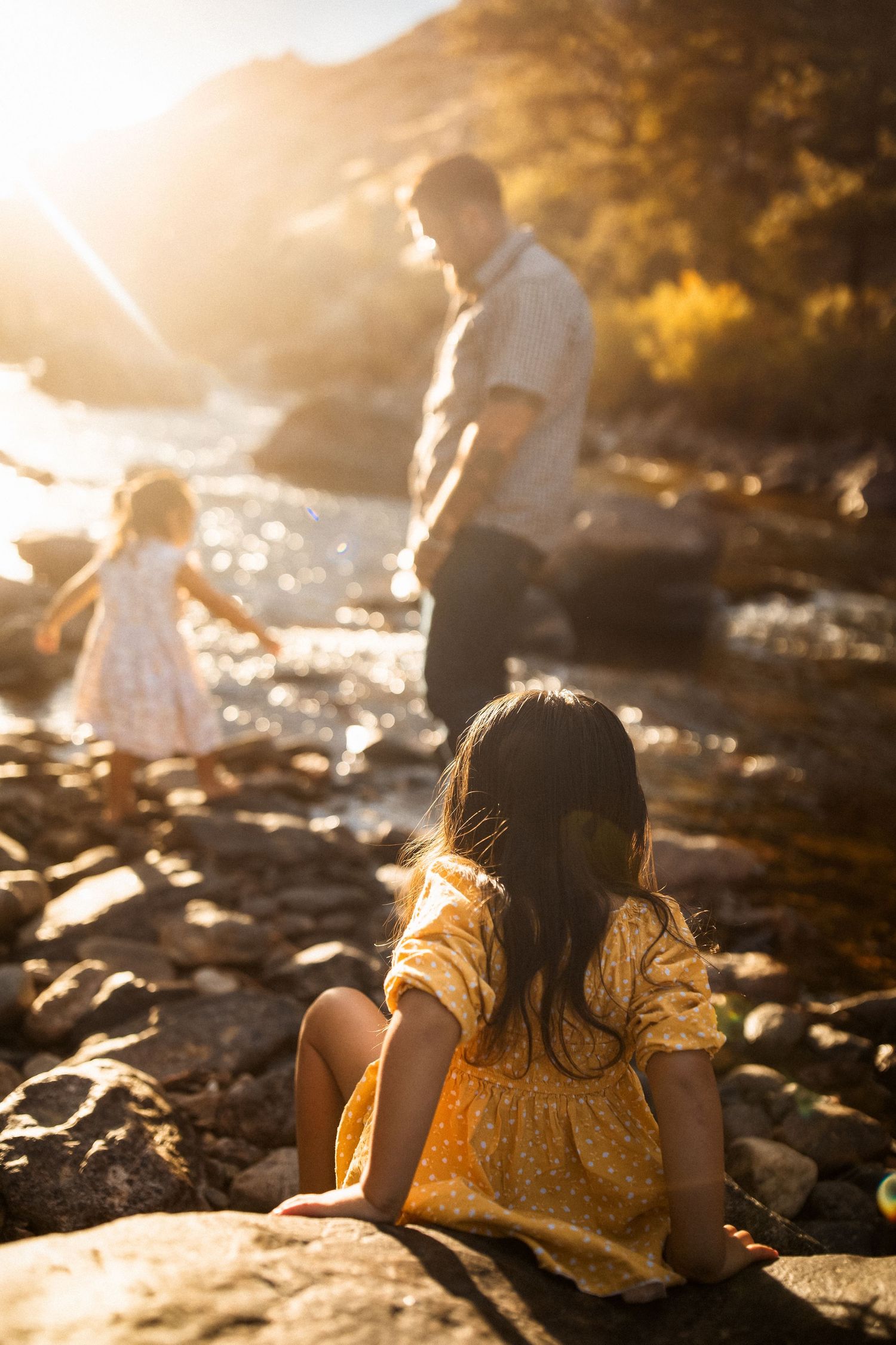 Family enjoying sunset by the water on rocky shore with golden light filtering through trees.