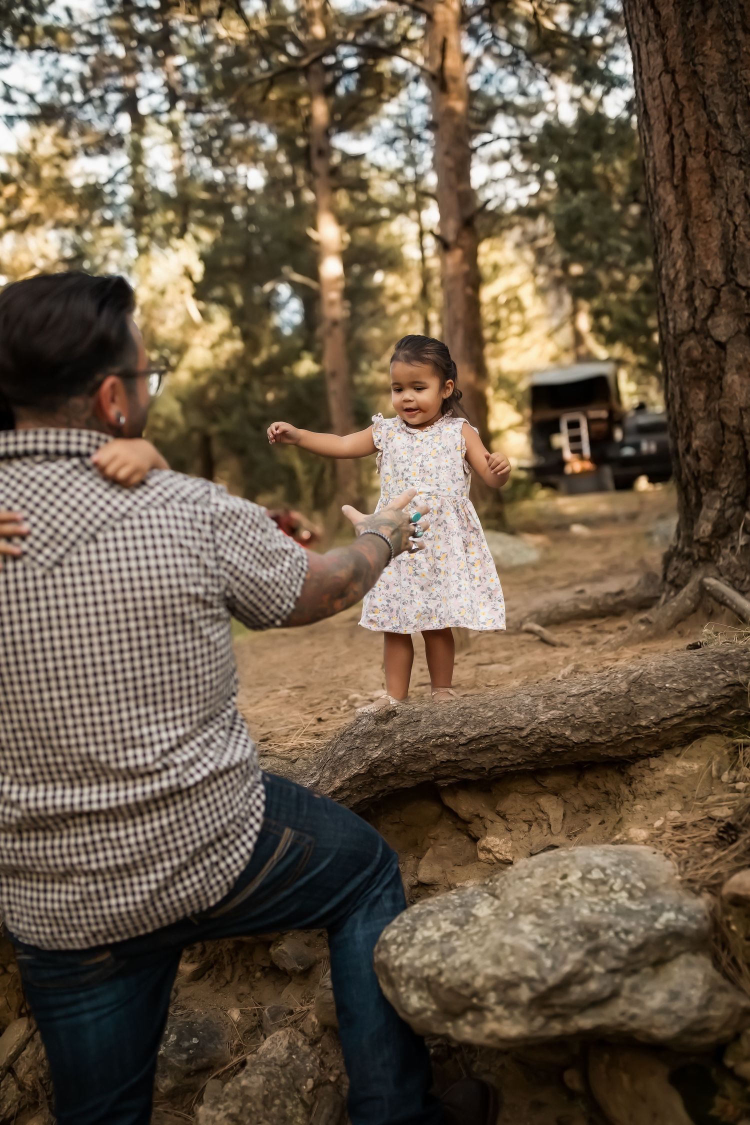 Child in white dress balancing on log while adult in plaid shirt assists in forest setting.