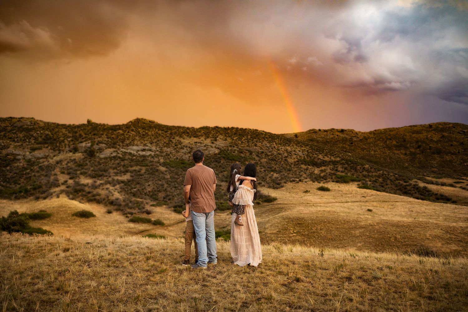 A beautiful rainbow arches over a desert mountain landscape at sunset with silhouettes in the foreground.