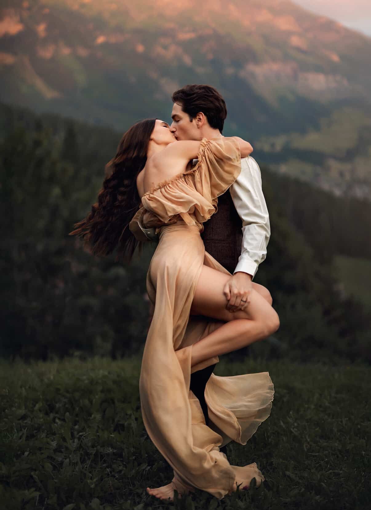 A couple shares a romantic kiss outdoors, man lifting woman in golden dress, mountains and greenery in background.