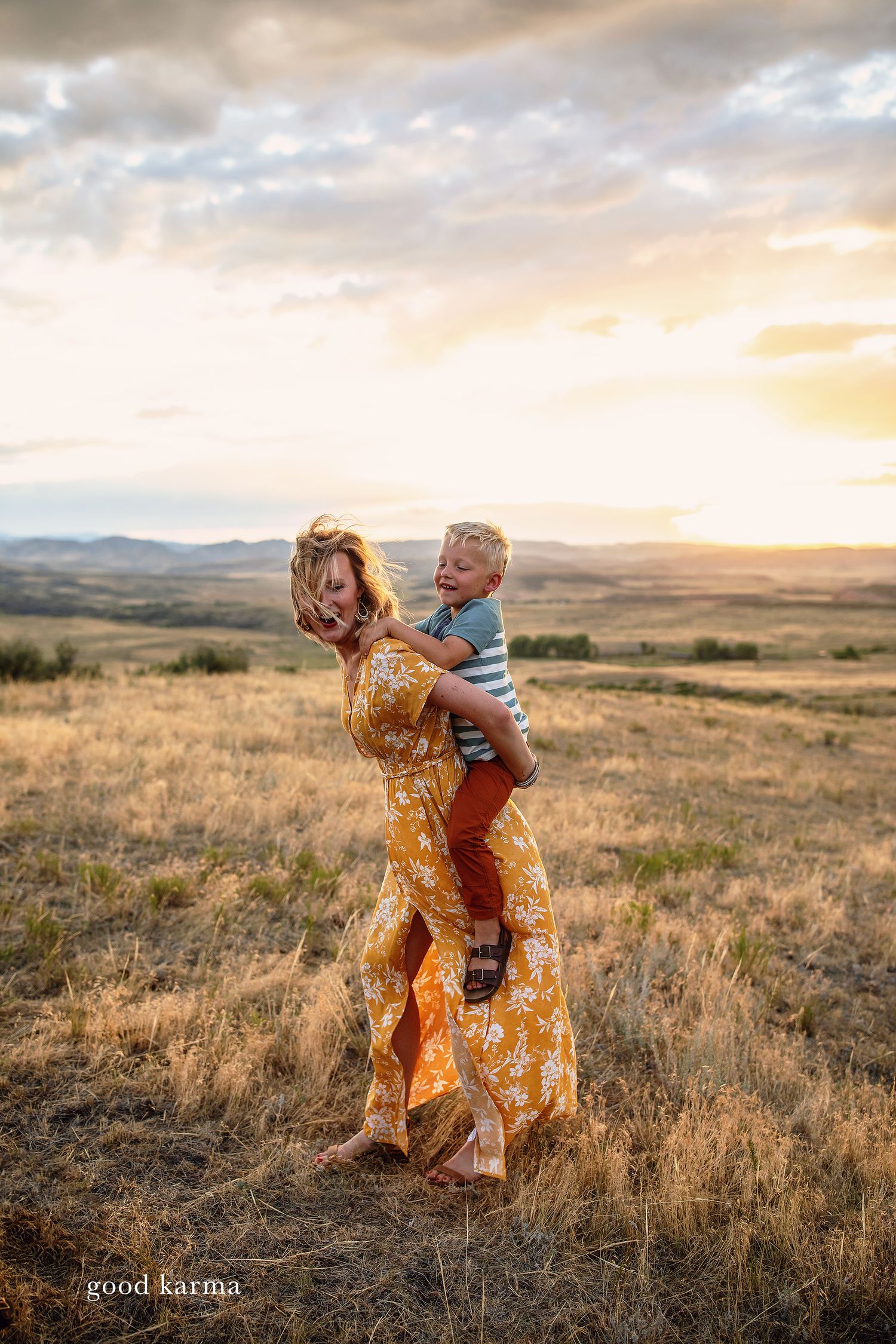 Two figures dance playfully in tall grass during golden hour with mountains on the horizon.