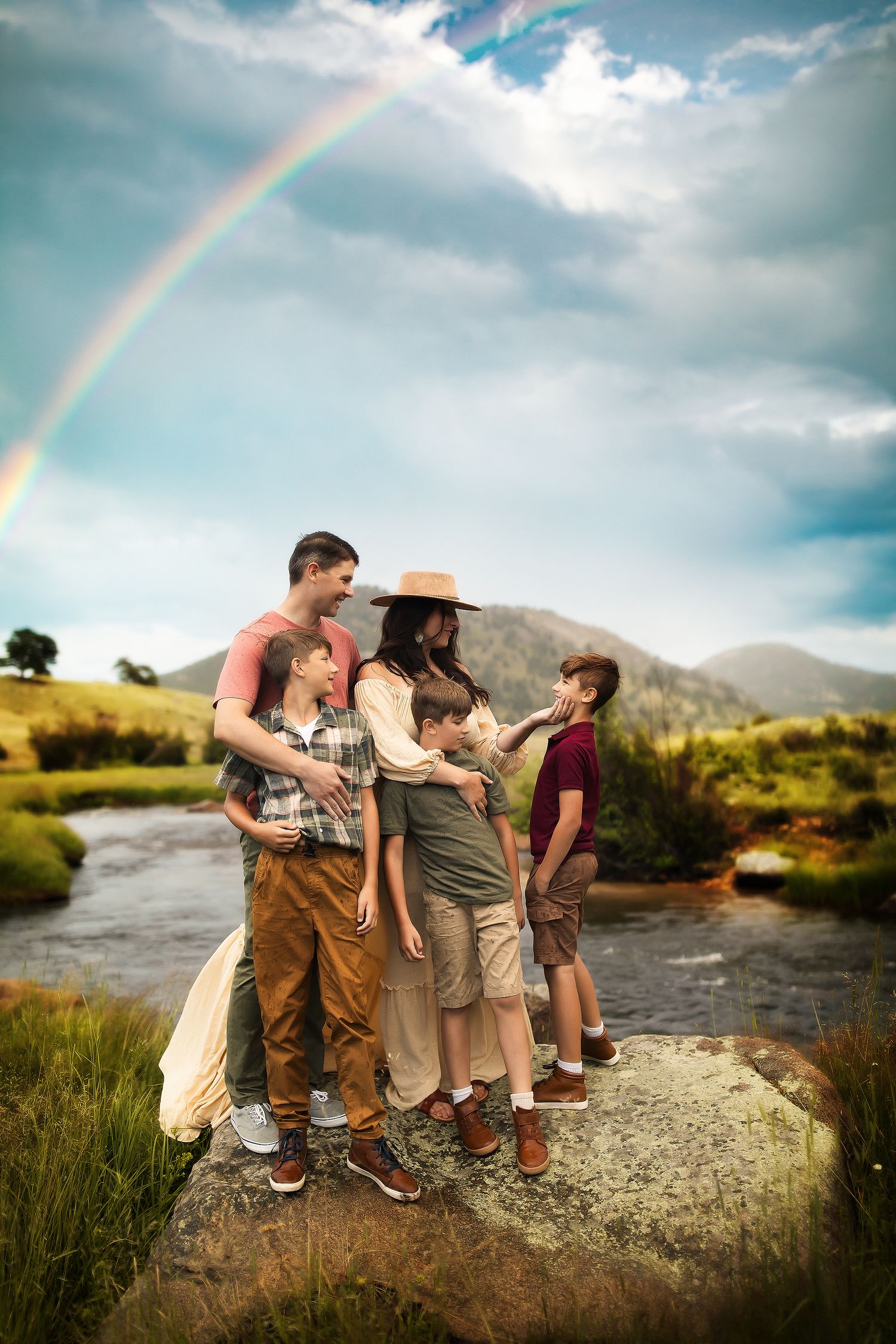 Group standing on a large rock by a river with a vibrant rainbow arching across the cloudy sky.