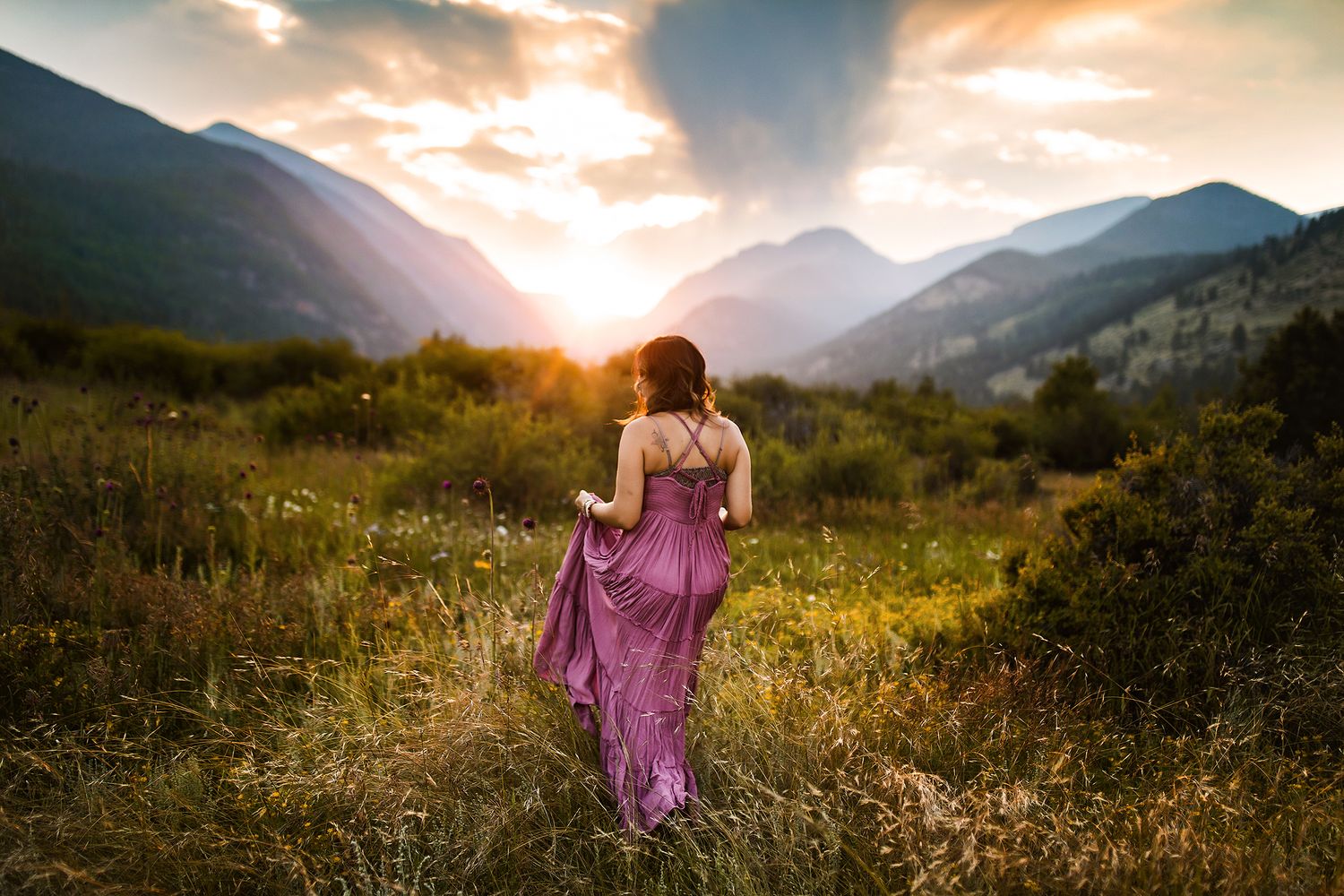 Flowing dress catches sunset light in mountain meadow with dramatic sky and wildflowers.