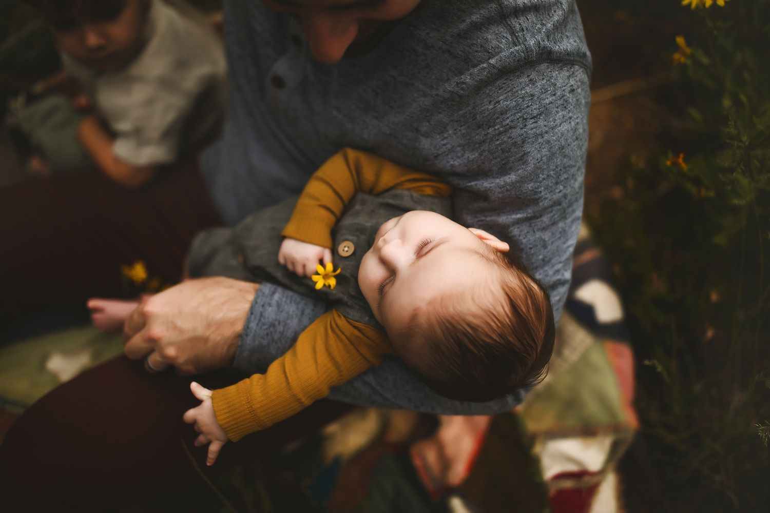 A small infant in a mustard yellow outfit being cradled in someone's arms outdoors.