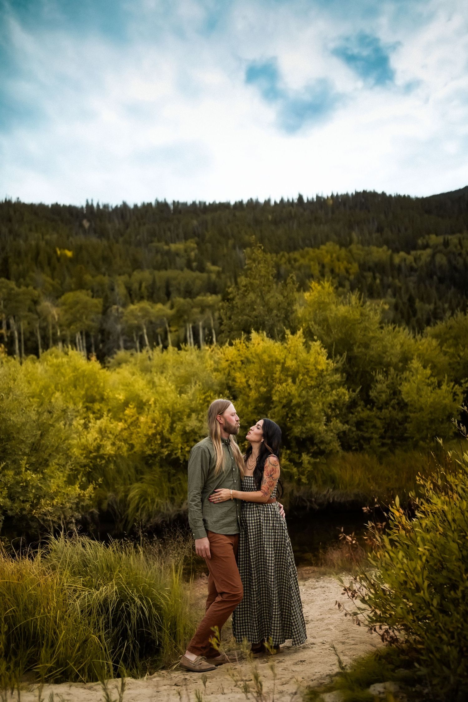 A couple embraces on a mountain trail surrounded by vibrant yellow foliage and evergreen forest under cloudy skies.