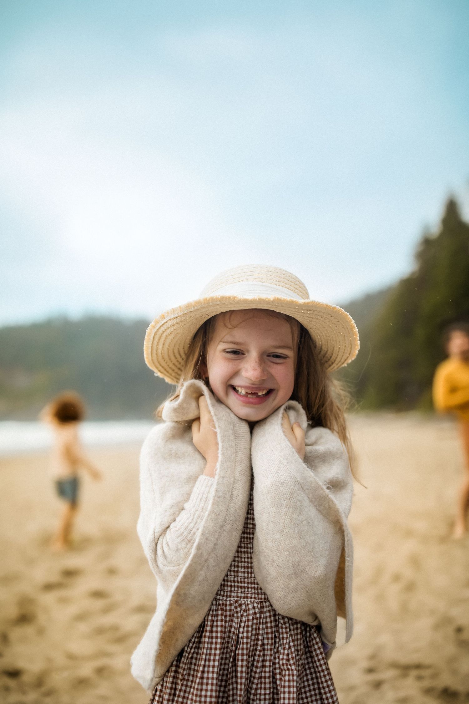 A child in a straw hat and checkered dress smiles joyfully while standing on a beach with others in the background.