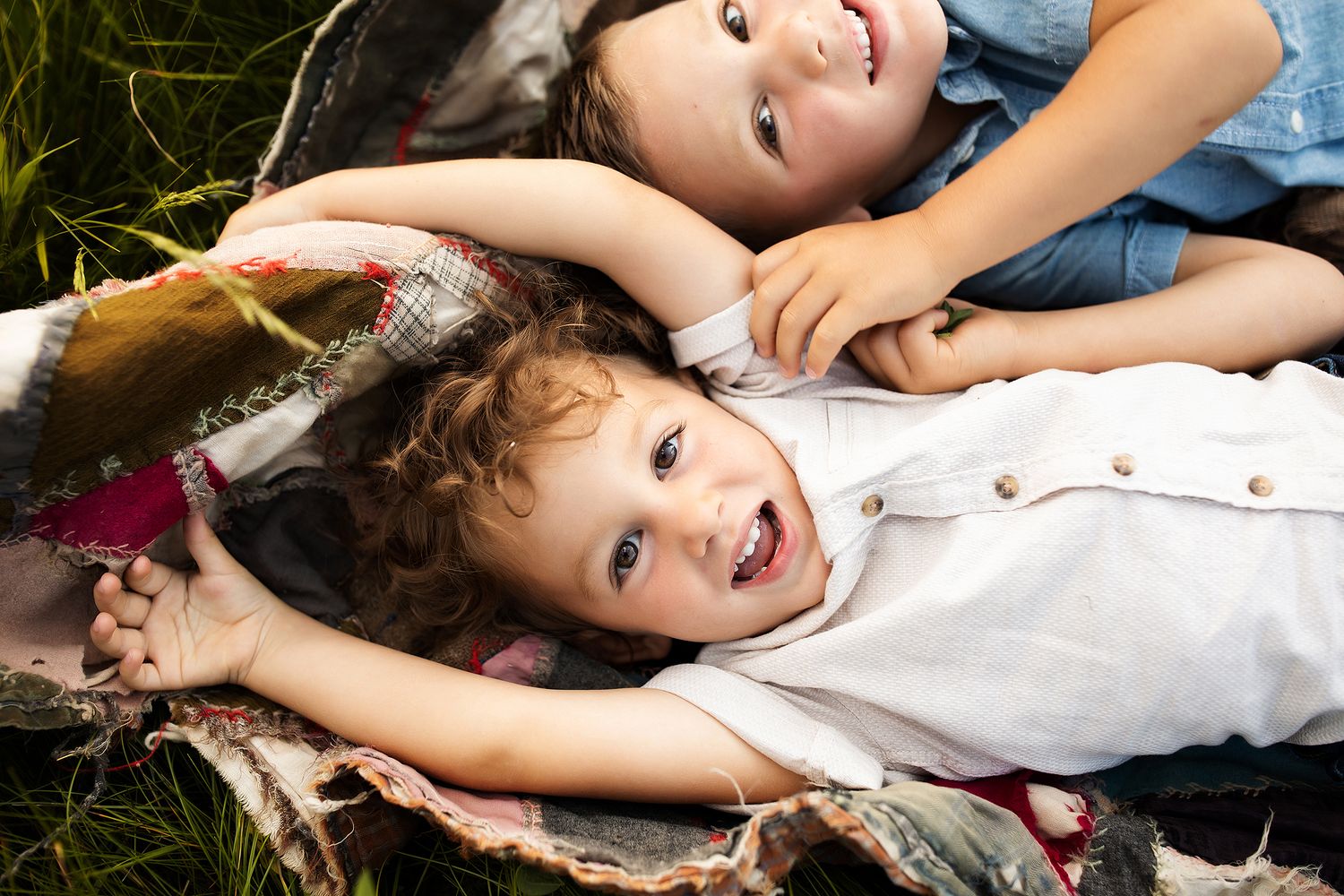 Two happy children lying on a blanket in the grass, smiling and laughing together outdoors.