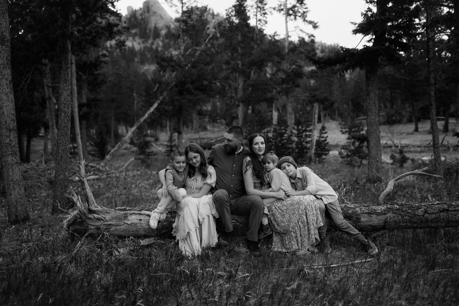 Black and white family portrait of five people sitting on a fallen log in a forest meadow.
