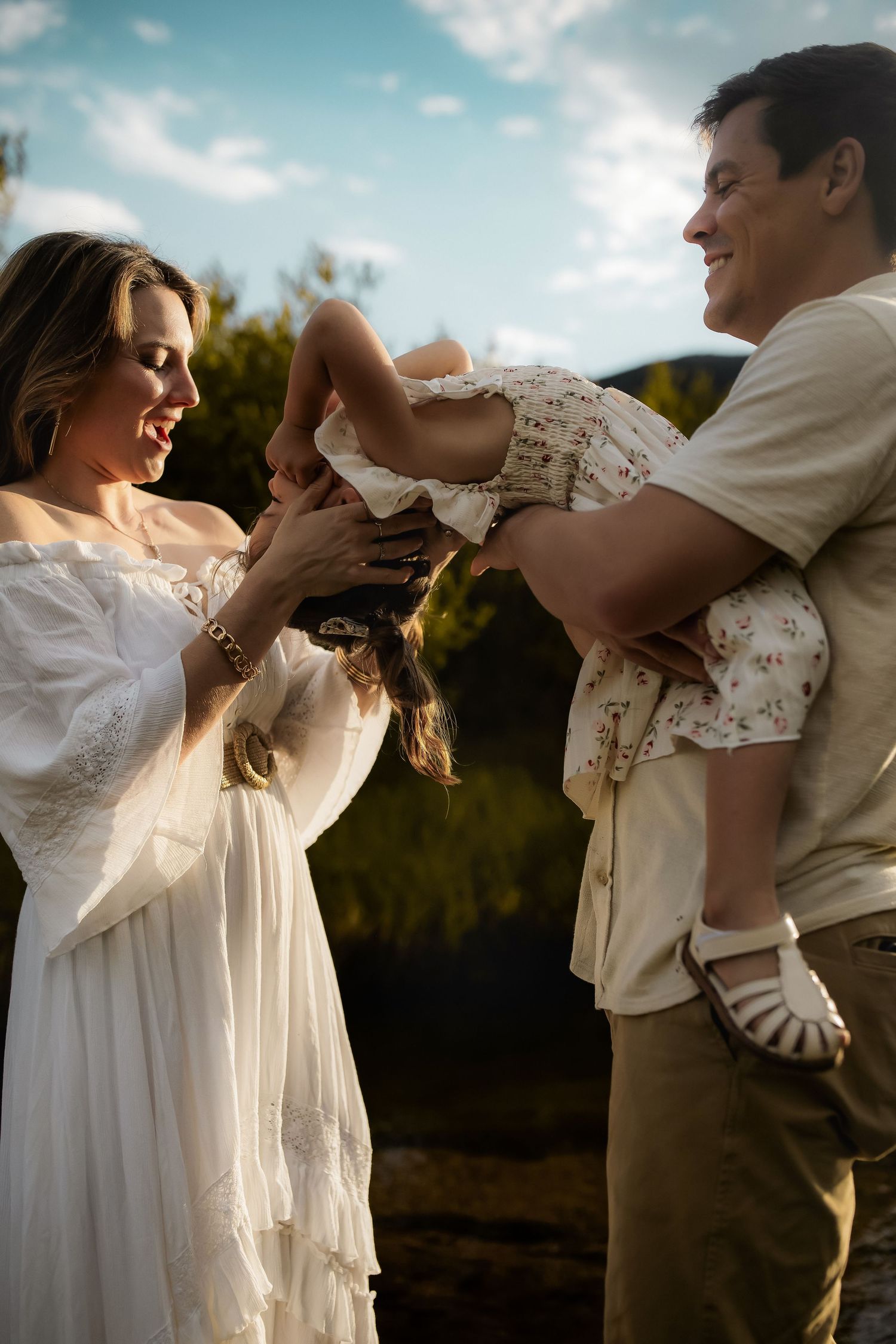 A happy family with a mother in white dress and father holding their young daughter upside down outdoors.