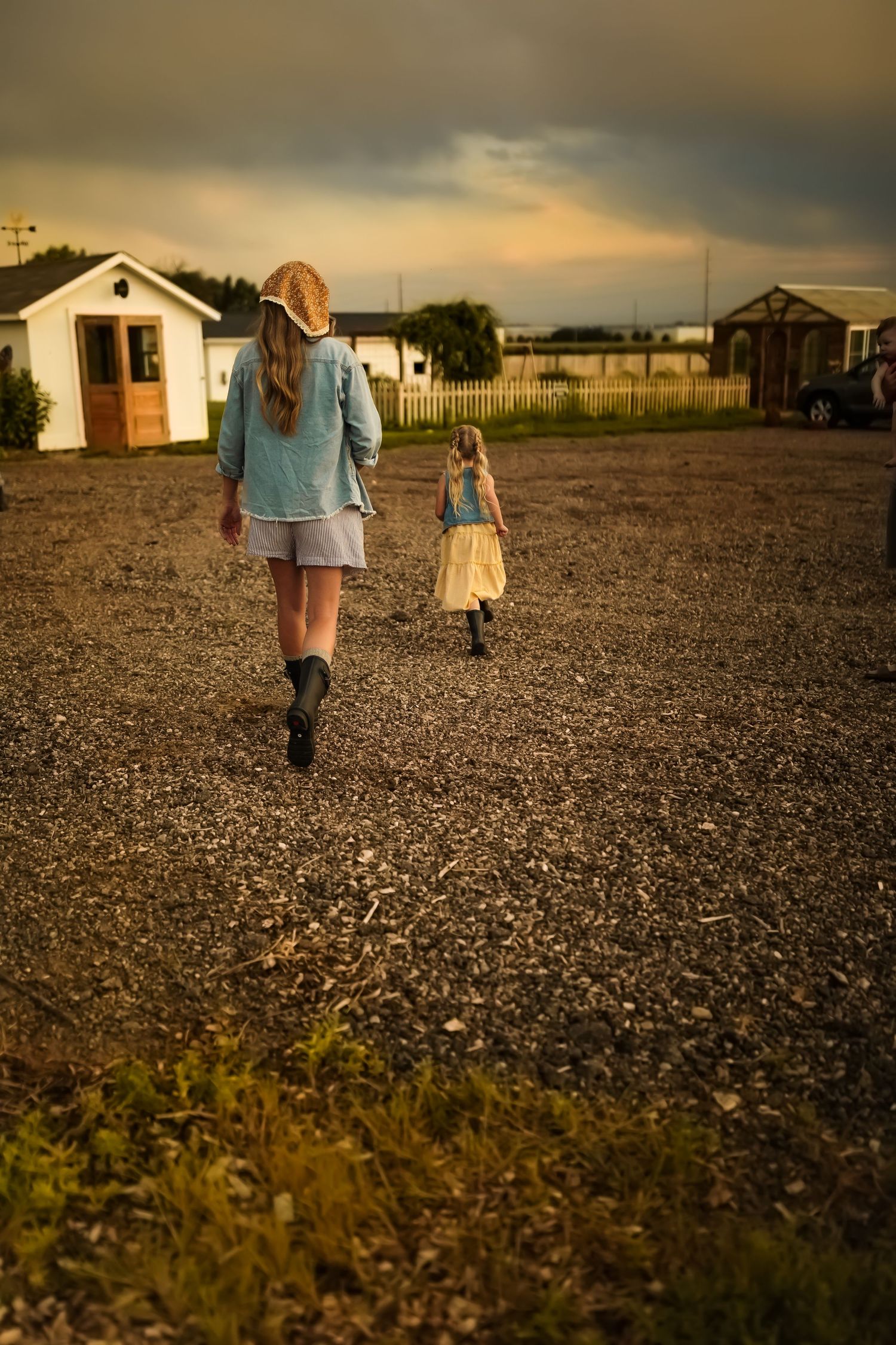 Two figures walk across a gravel lot at sunset with rural buildings visible in the background.