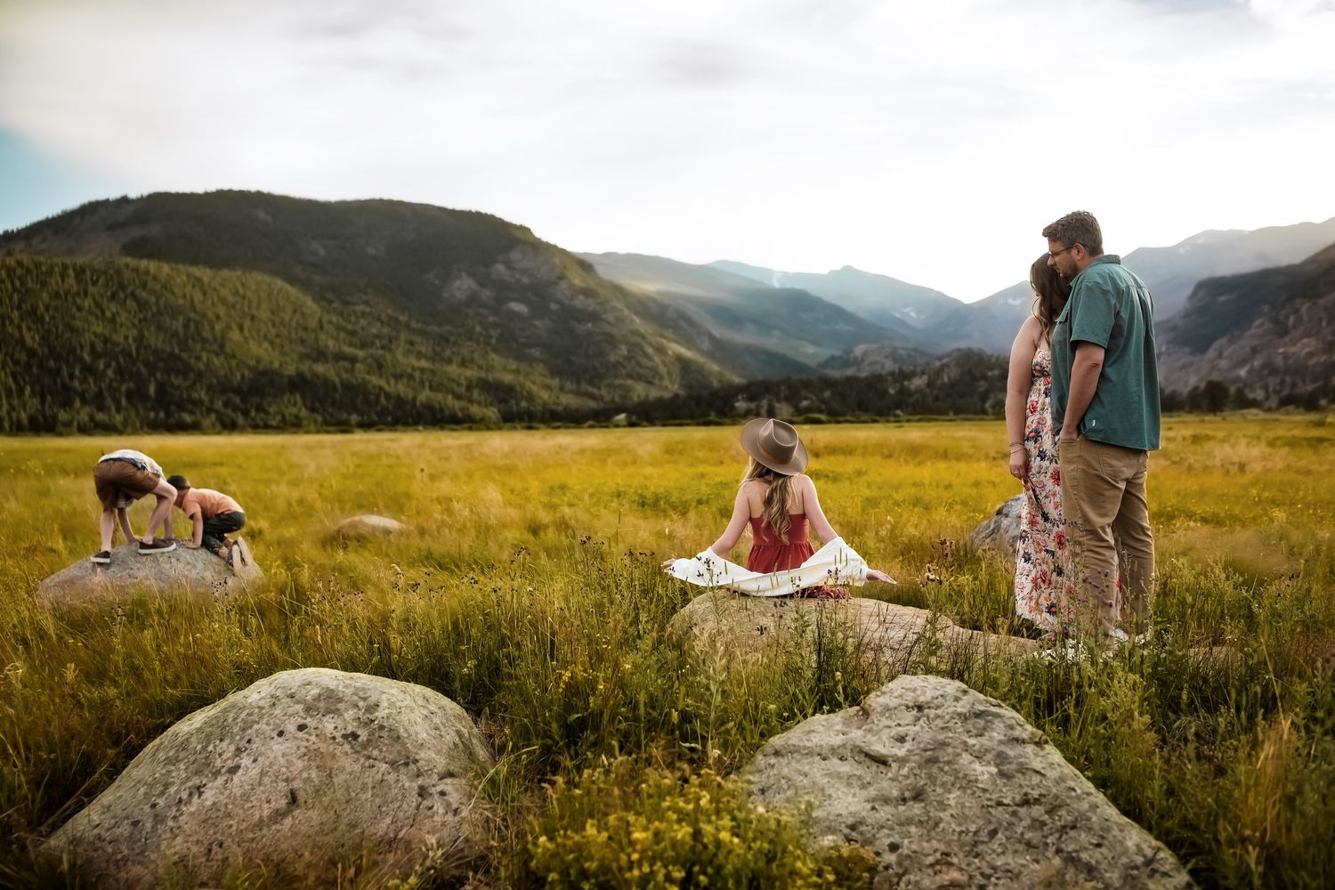 A group of people enjoy a scenic mountain meadow view while sitting on large boulders at sunset.